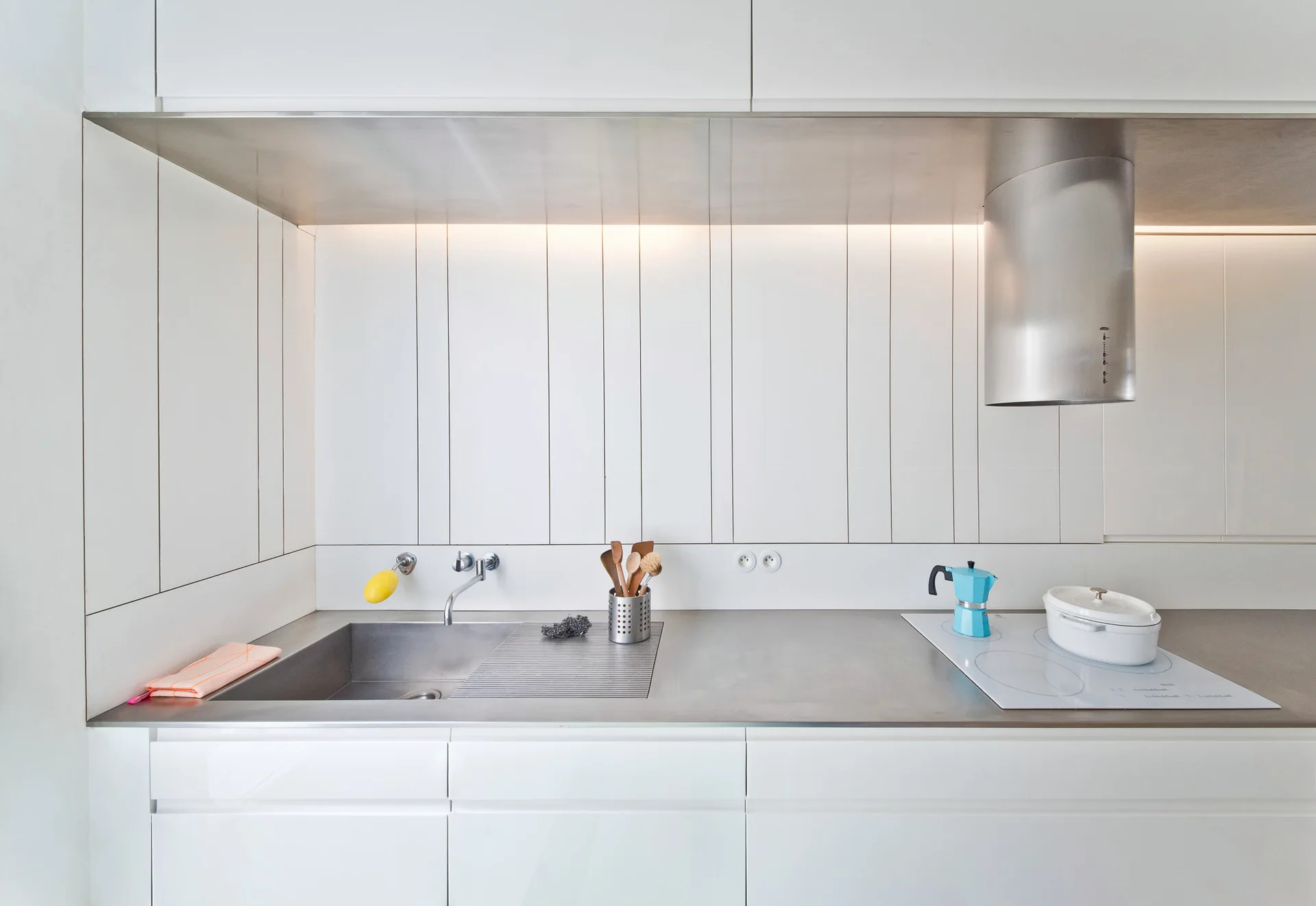 A modern kitchen workspace with white flat-panel cabinets, a stainless steel countertop with an integrated sink, and a vertically paneled backsplash.