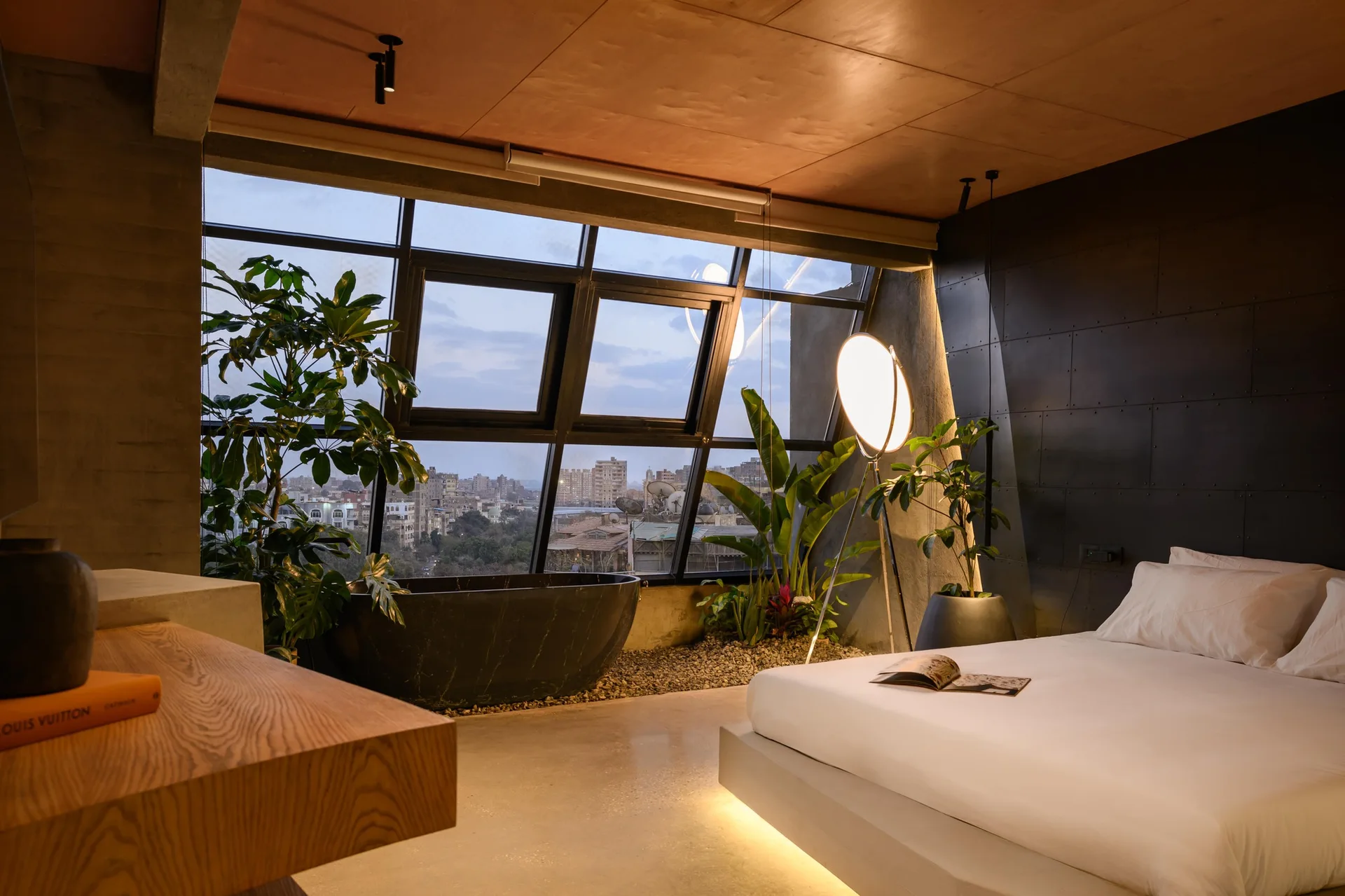An industrial bedroom featuring a low platform bed with under-bed lighting, a black freestanding bathtub, and a large slanted window.