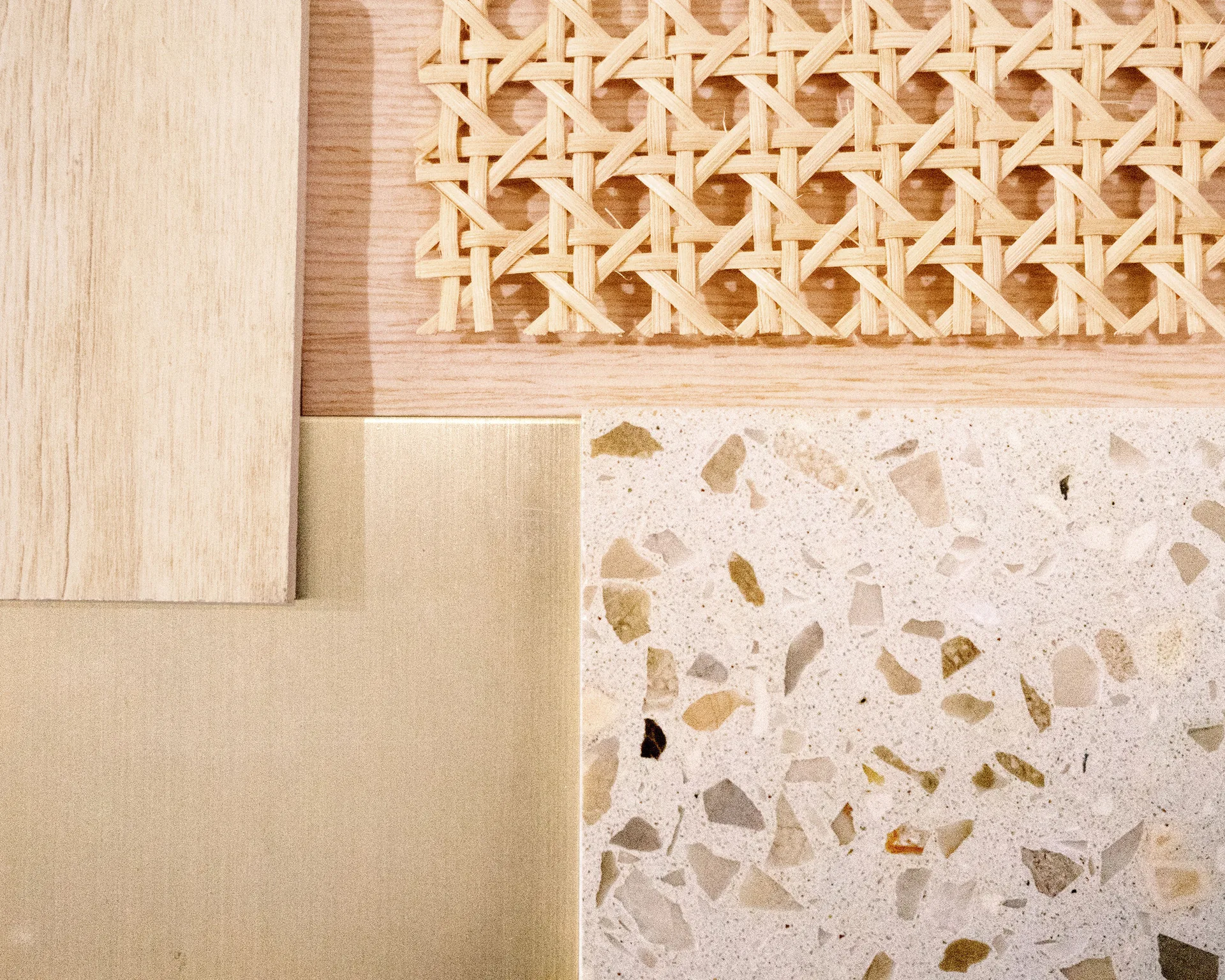 A top-down view of design material samples including woven cane webbing, light wood veneer, brushed brass, and a speckled terrazzo tile.