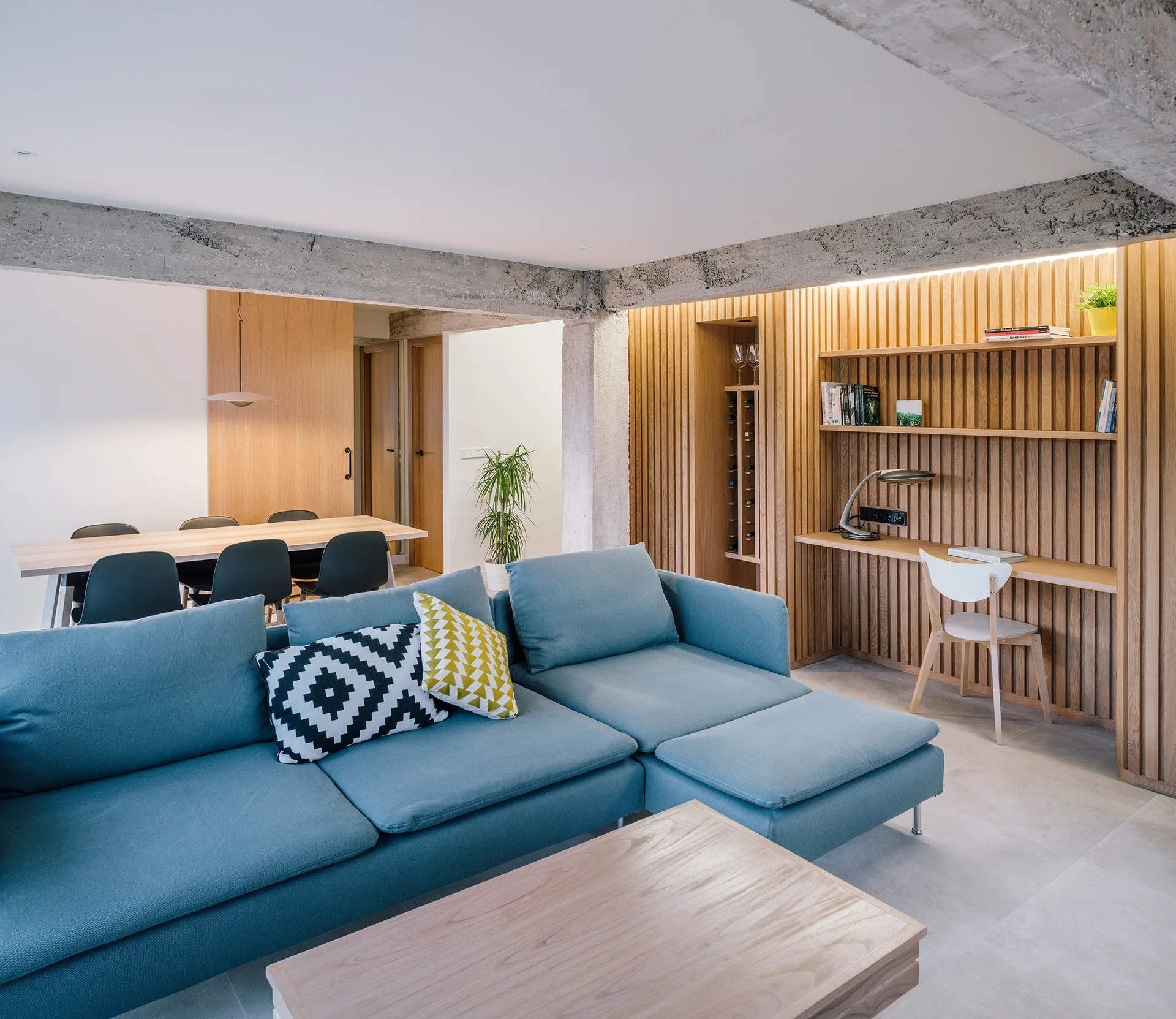 A modern open-plan living room featuring a blue sectional sofa, a built-in wooden desk integrated into a slatted wood wall, and exposed concrete ceiling beams.
