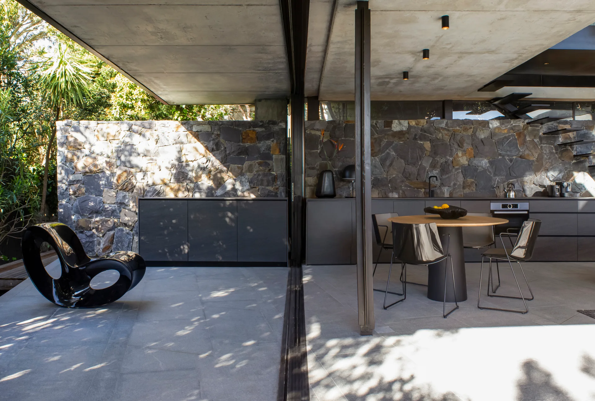 A contemporary indoor-outdoor patio features a grey stone wall, charcoal kitchen cabinetry, a round wooden dining table, and a sculptural black chair.