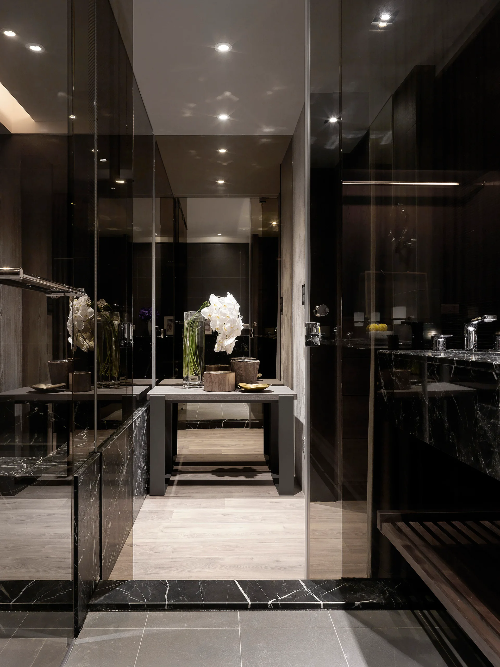 A modern dark bathroom features a black veined marble vanity, glass partitions, mirrored walls, and a grey console table holding white orchids.