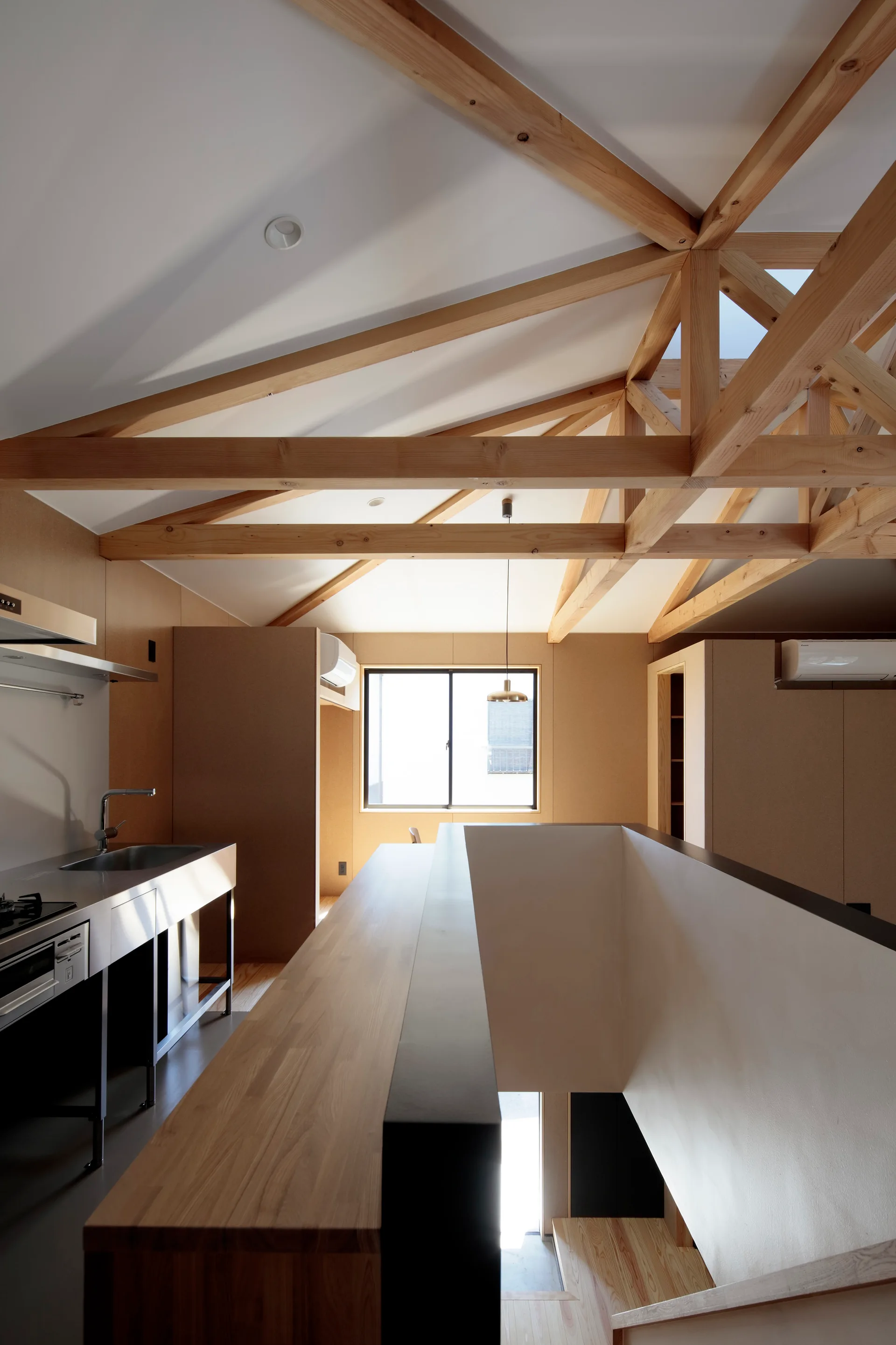A minimalist loft kitchen featuring exposed timber roof trusses, a wooden counter bordering a staircase void, and light wood wall panels.