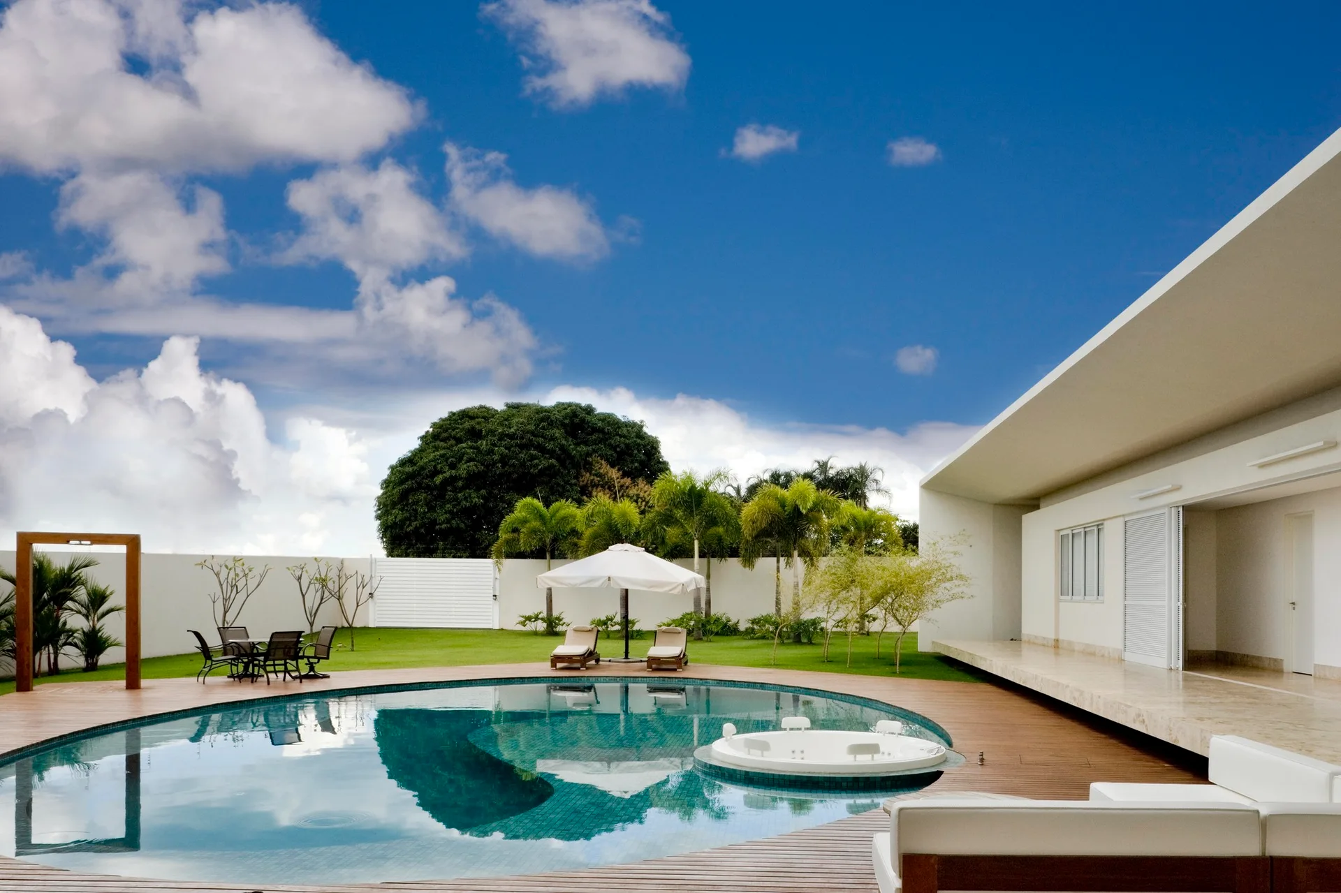 A curved backyard swimming pool with an integrated circular hot tub is surrounded by a wooden plank deck, lawn, palm trees, and a white modern house.