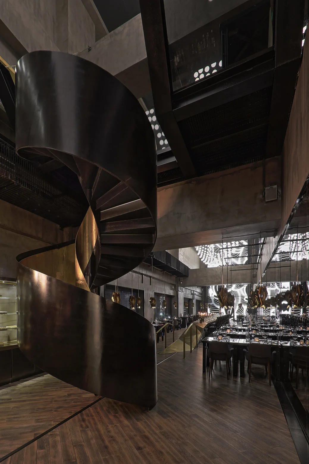 A dark metal spiral staircase with a brass interior stands in a spacious industrial restaurant dining hall with wooden floors.