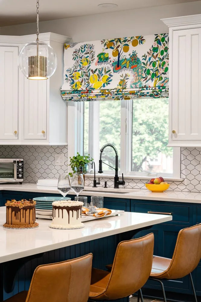 A residential kitchen featuring navy blue lower cabinetry, white upper cabinets, a hexagonal tile backsplash, and a botanical print Roman shade.