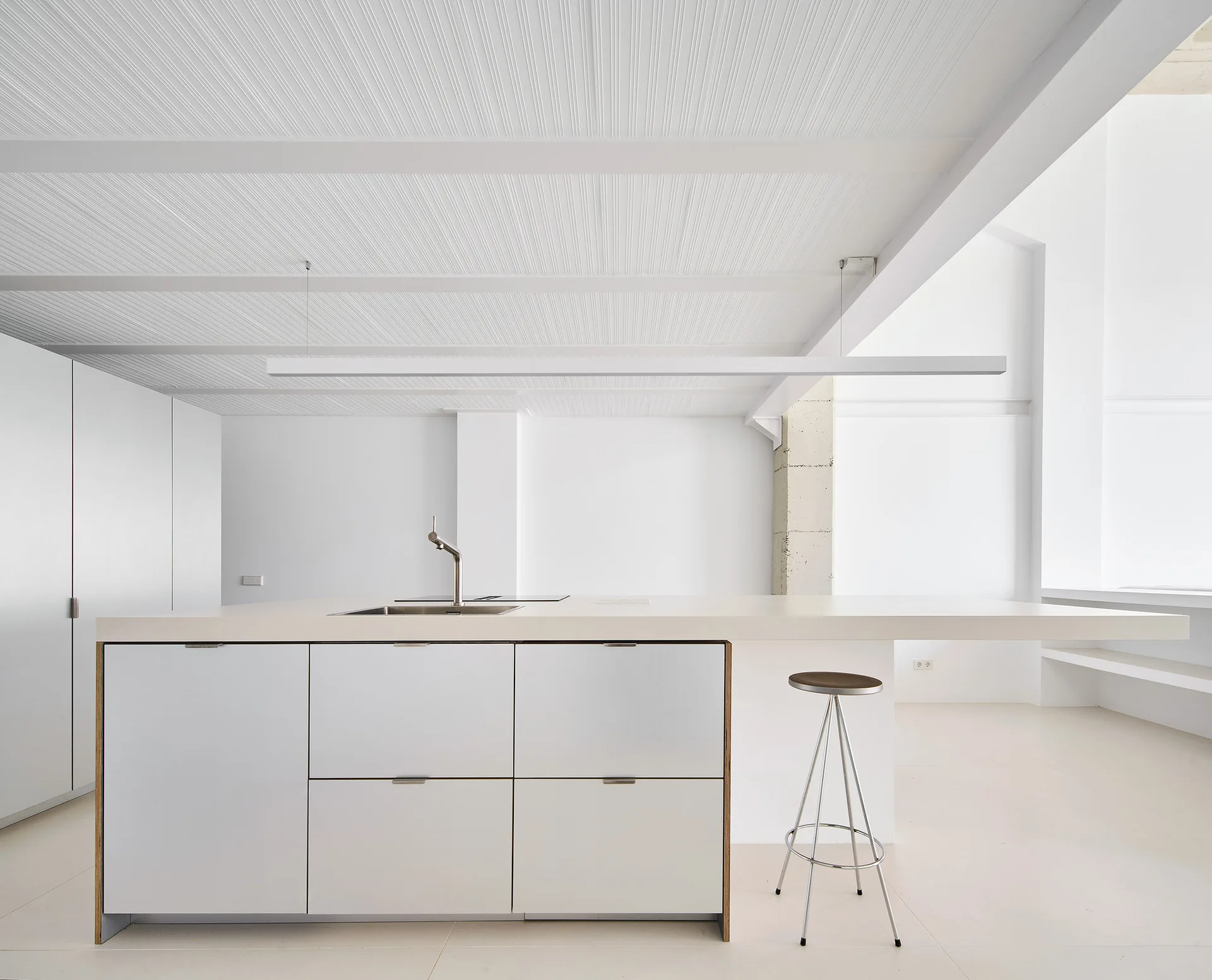 A minimalist white kitchen features a central island with a sleek faucet, a single metal stool, a linear pendant light, and a ribbed ceiling.