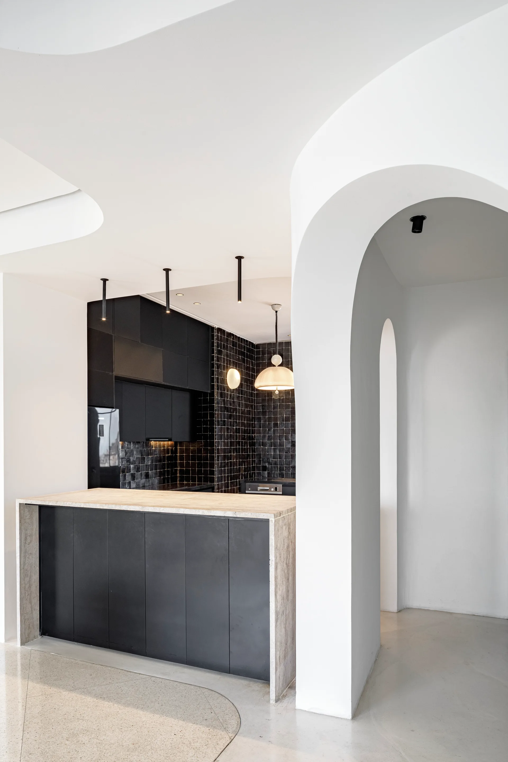 A modern kitchen featuring a matte black cabinetry wall, black tile backsplash, stone island, and a distinctive white curved plaster archway.