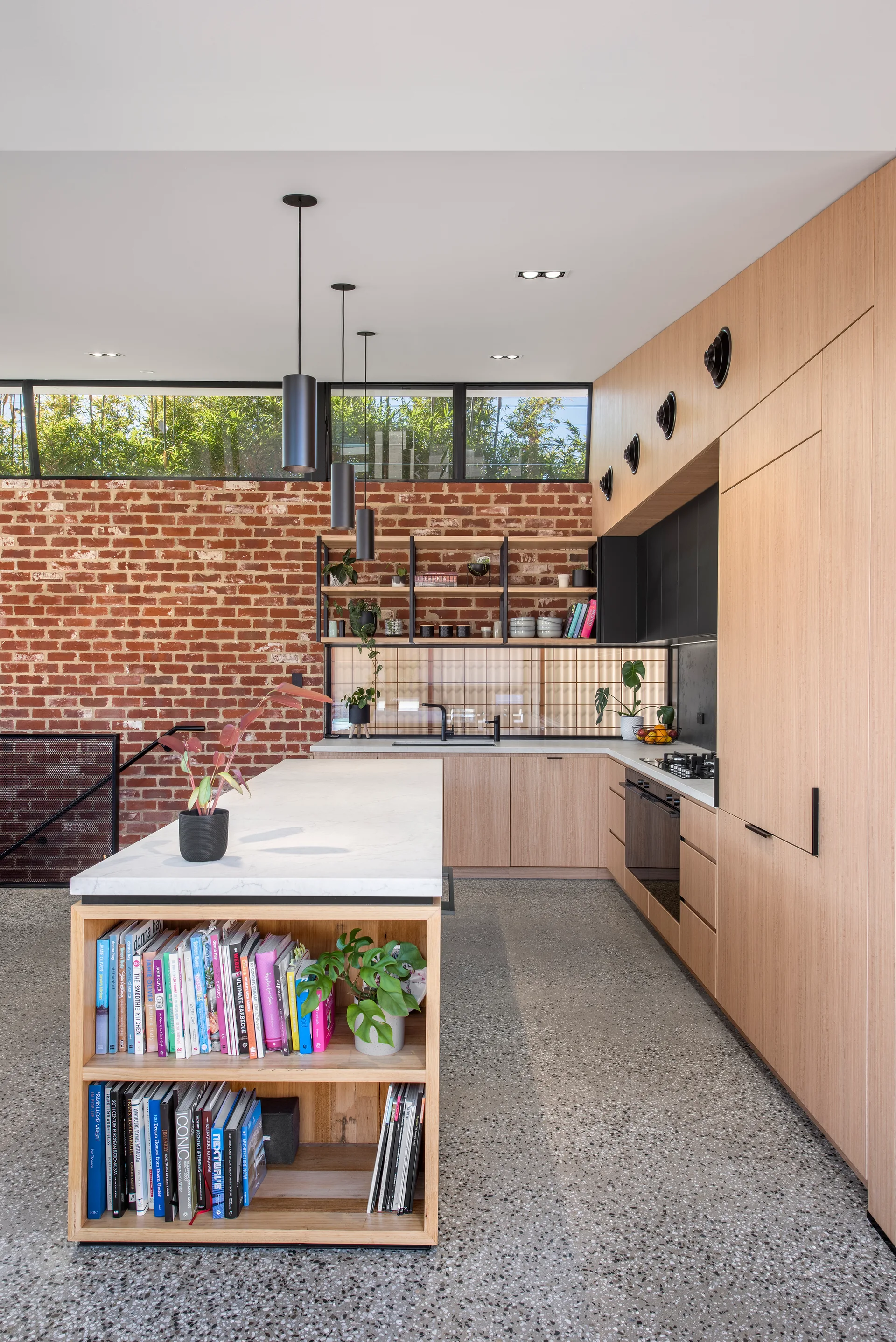 A contemporary kitchen features a wood island with built-in bookshelves, flat-front wood cabinets, an exposed brick wall, and grey terrazzo floors.