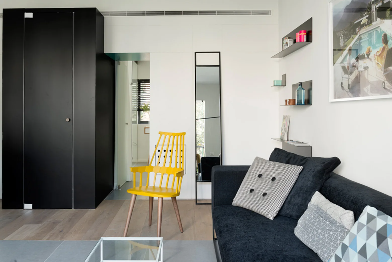 A modern living room features a dark fabric sofa, a vibrant yellow spindle chair, a tall black cabinet, and slim metal floating shelves.