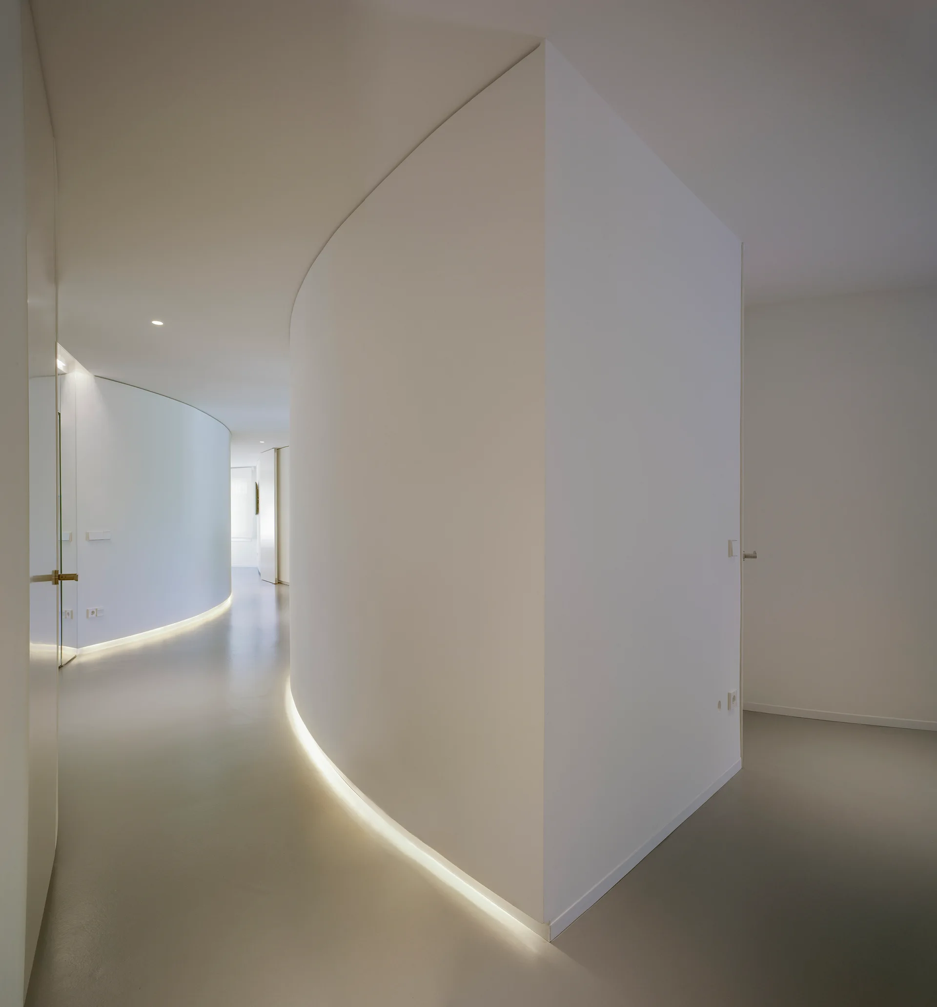 A minimalist white hallway featuring a sweeping curved wall illuminated by a strip of warm recessed lighting along the base of the floor.