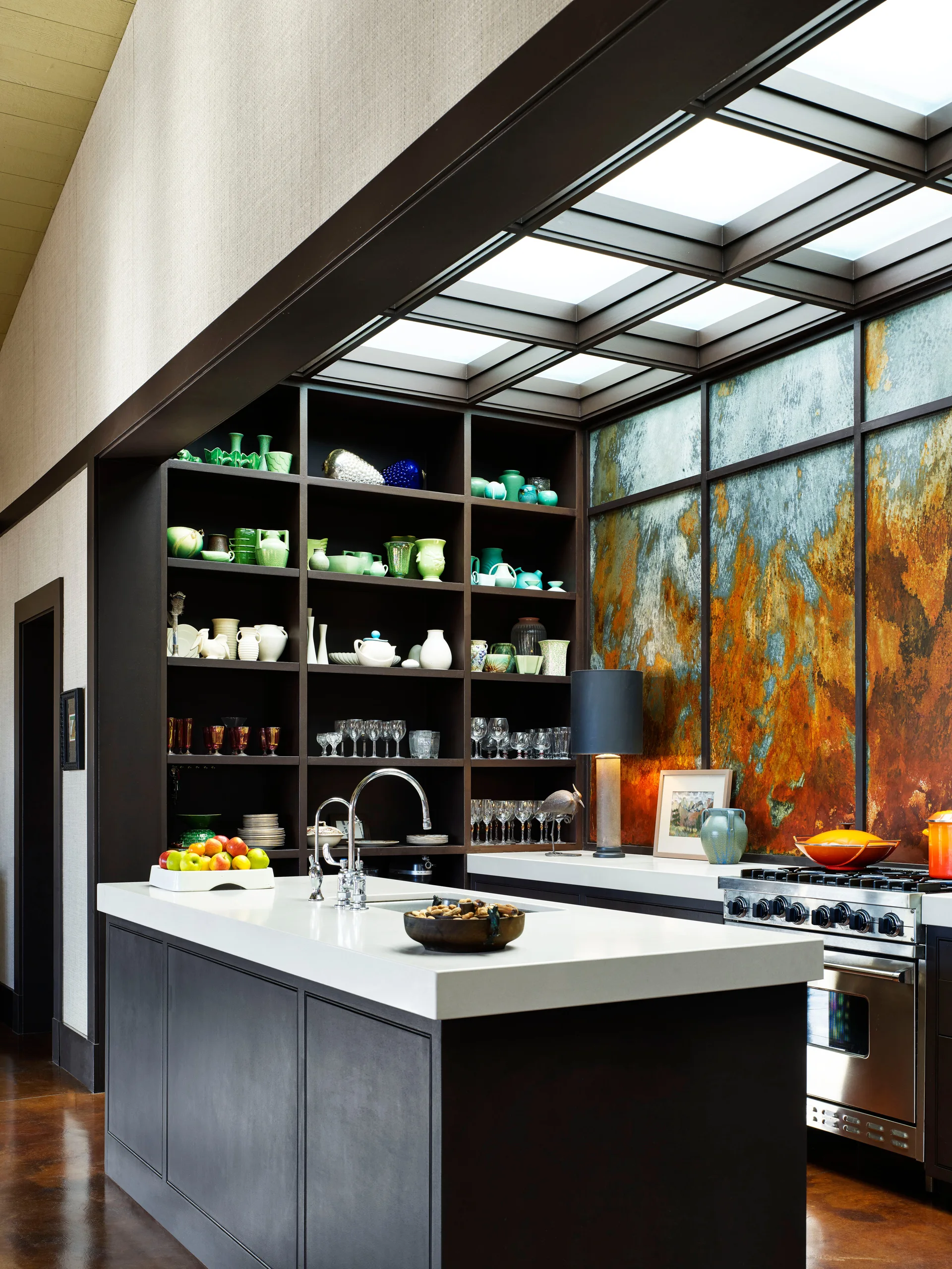 A dark kitchen island sits beneath a grid skylight, alongside a rusted metal backsplash and open shelves holding green and white ceramic dishware.