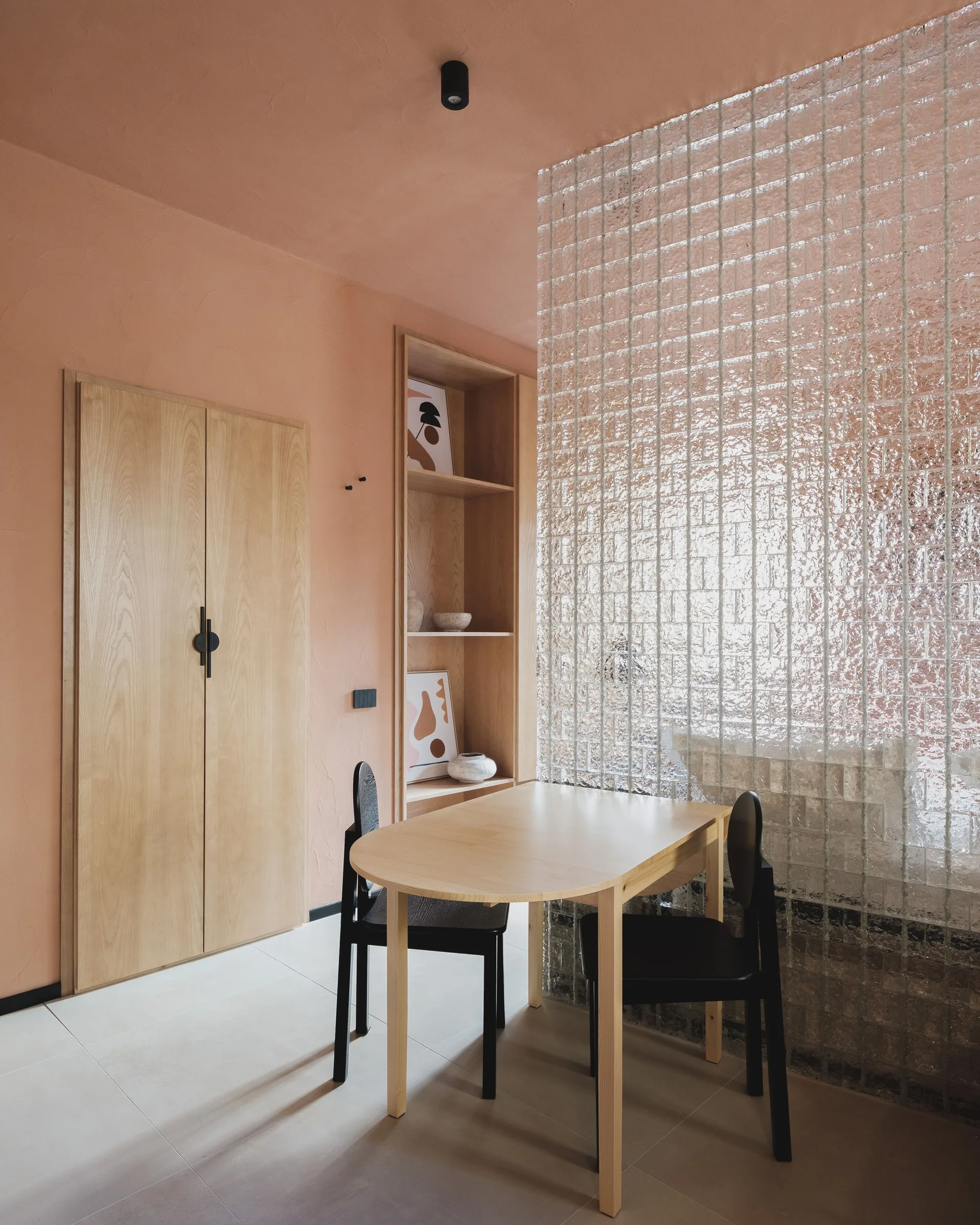 A minimalist dining area with a light wood table, black chairs, a glass block partition, and peach-colored walls.