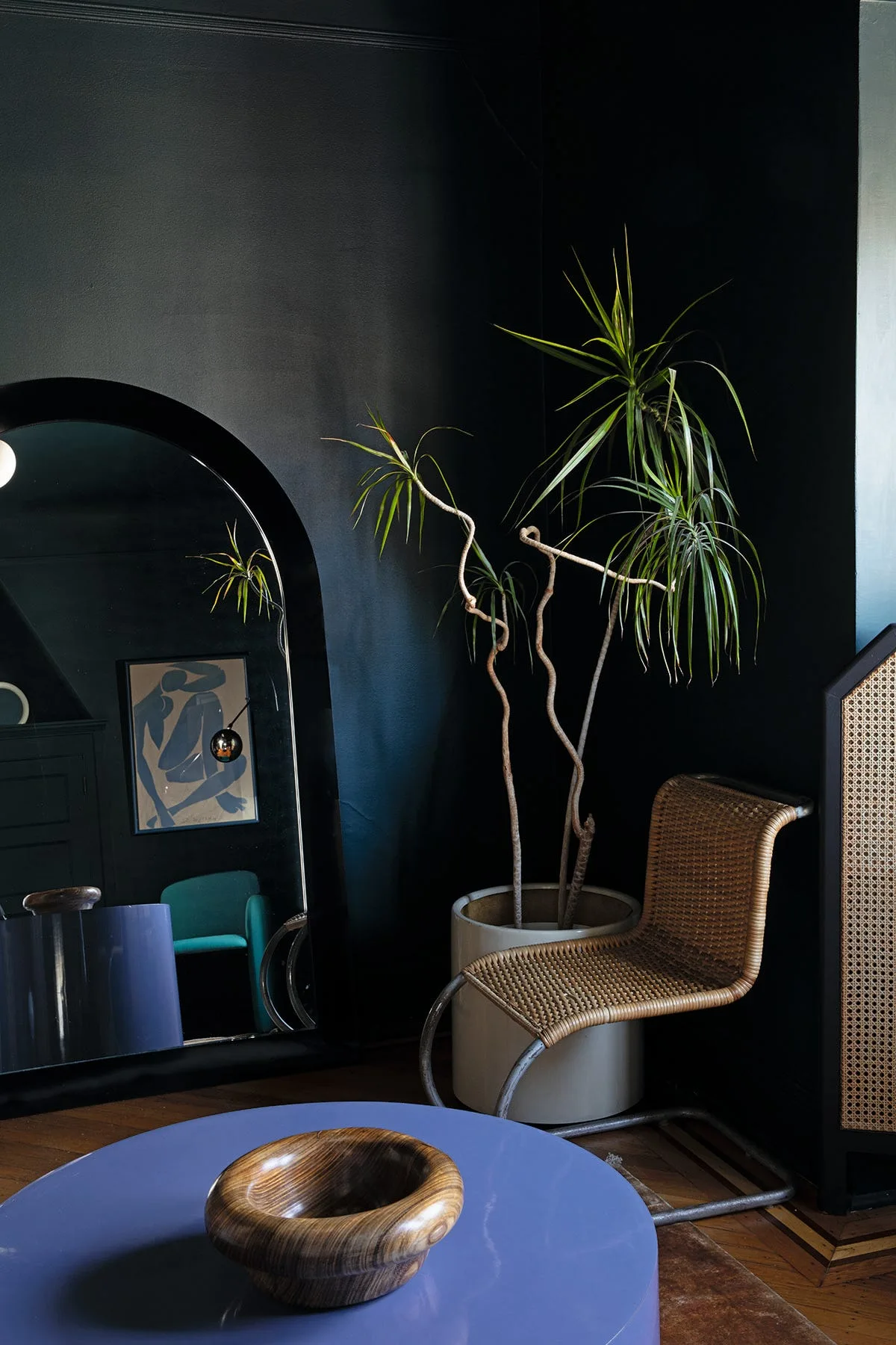 A corner vignette with matte charcoal walls features a large arched floor mirror, a tall potted plant, and a wicker chair on a wood floor.