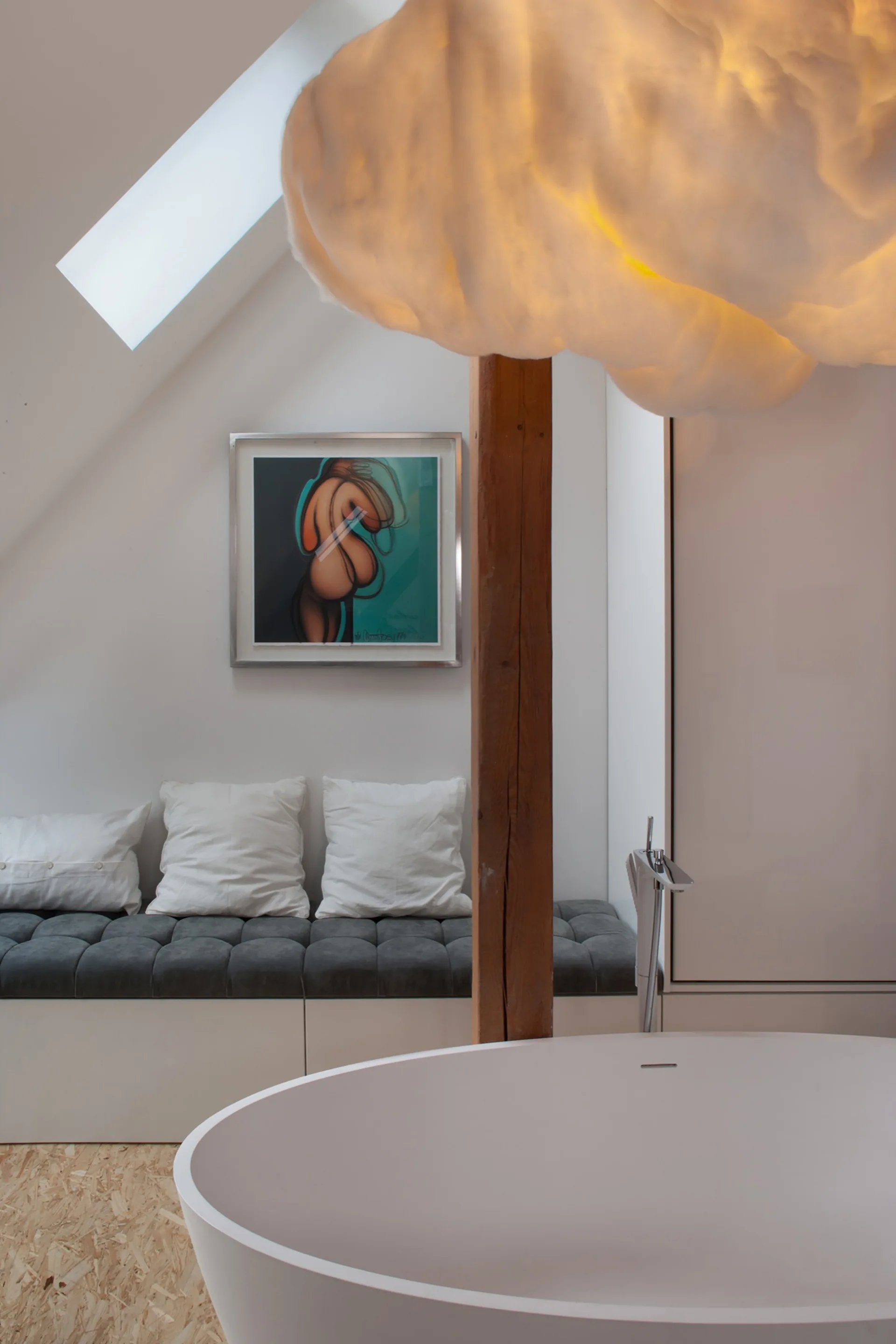A modern attic bathroom featuring a white freestanding bathtub beneath a cloud-shaped pendant light and a grey tufted bench under a skylight.