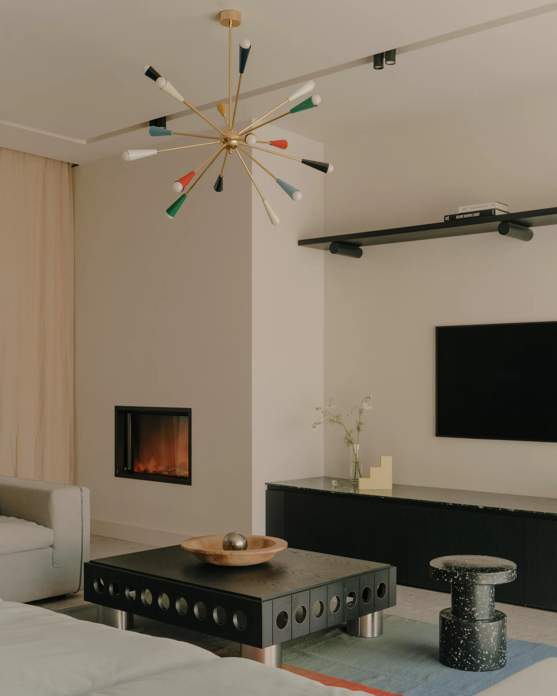 A modern living room features a recessed wall fireplace, a low black coffee table, a speckled stool, and a brass sputnik chandelier.