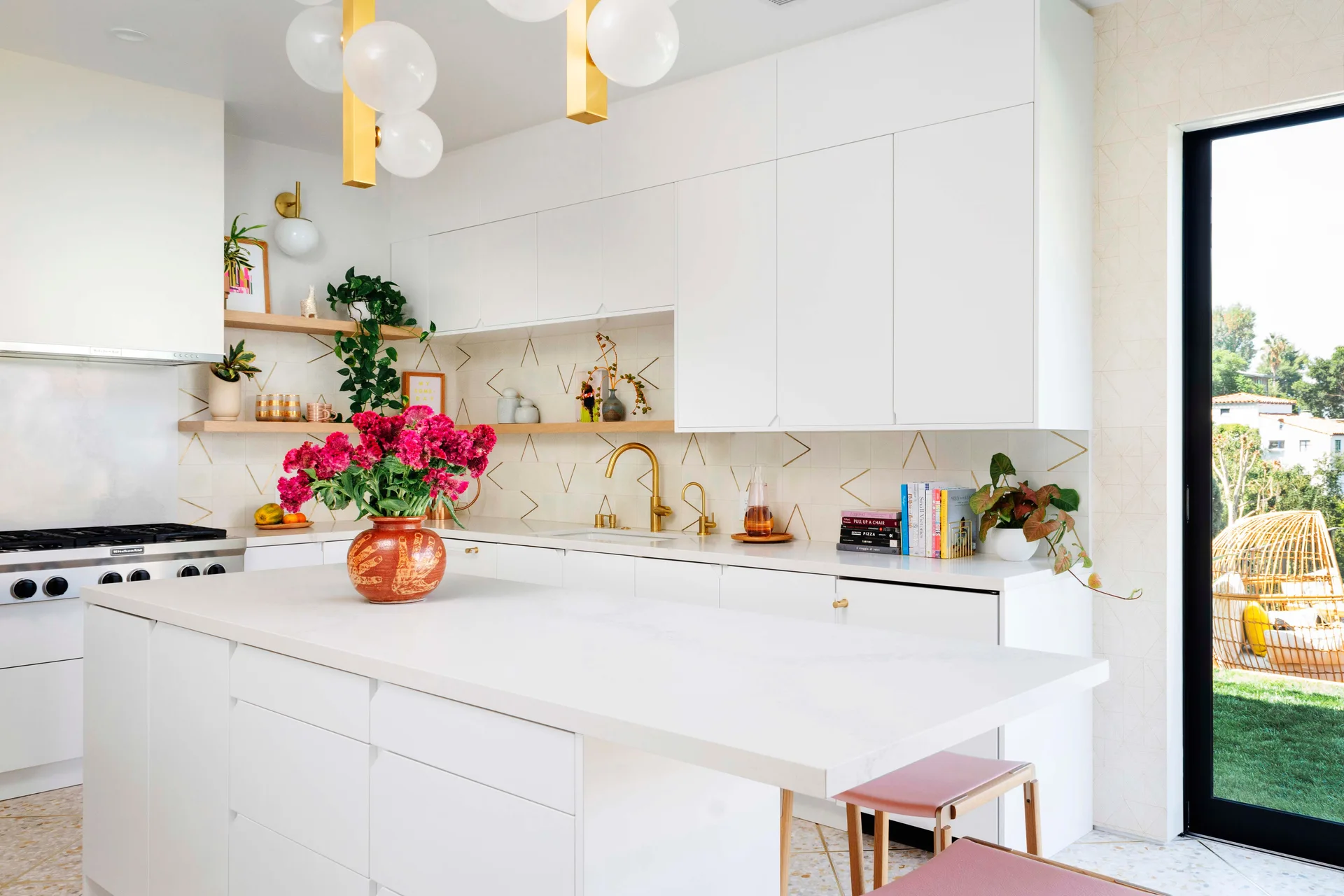 A bright white kitchen features a large central island, a gold geometric patterned backsplash, and modern globe pendant lights.