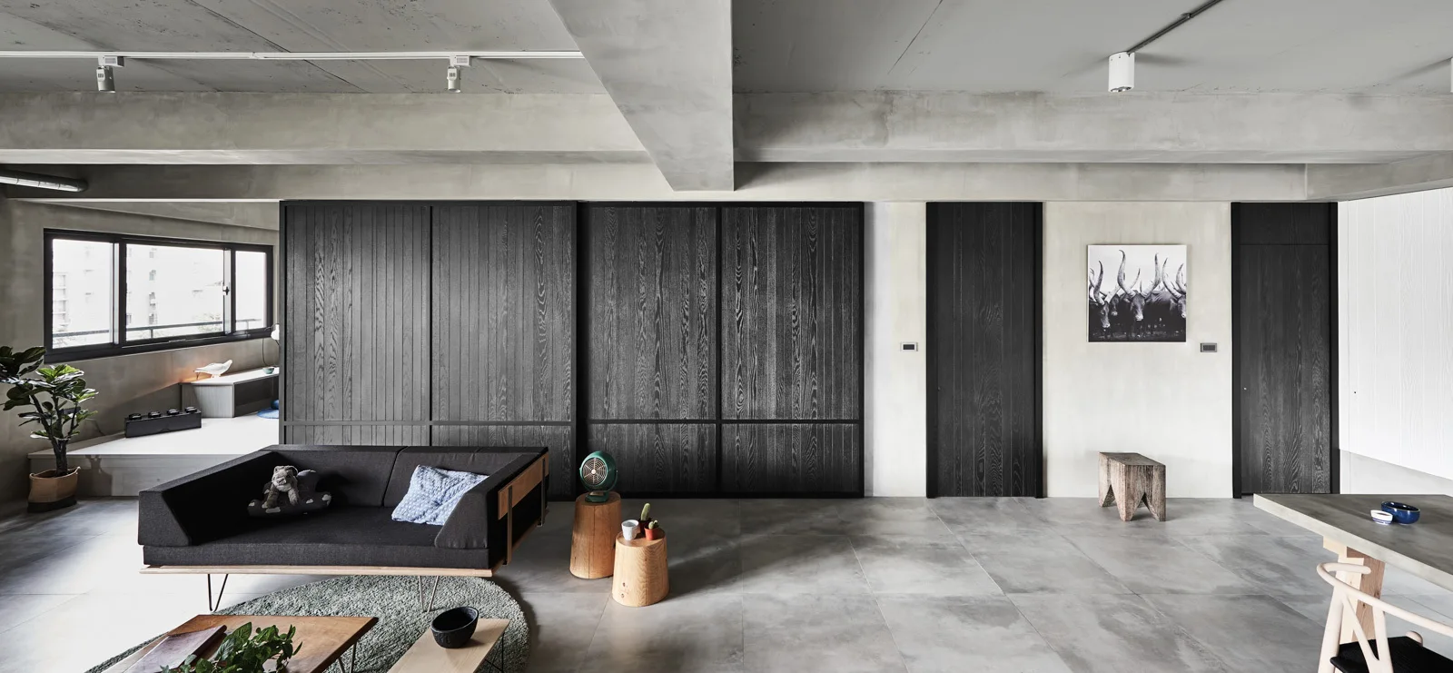 A minimalist living room featuring an exposed concrete ceiling, a low black sofa, and large charcoal wood sliding doors on the wall.