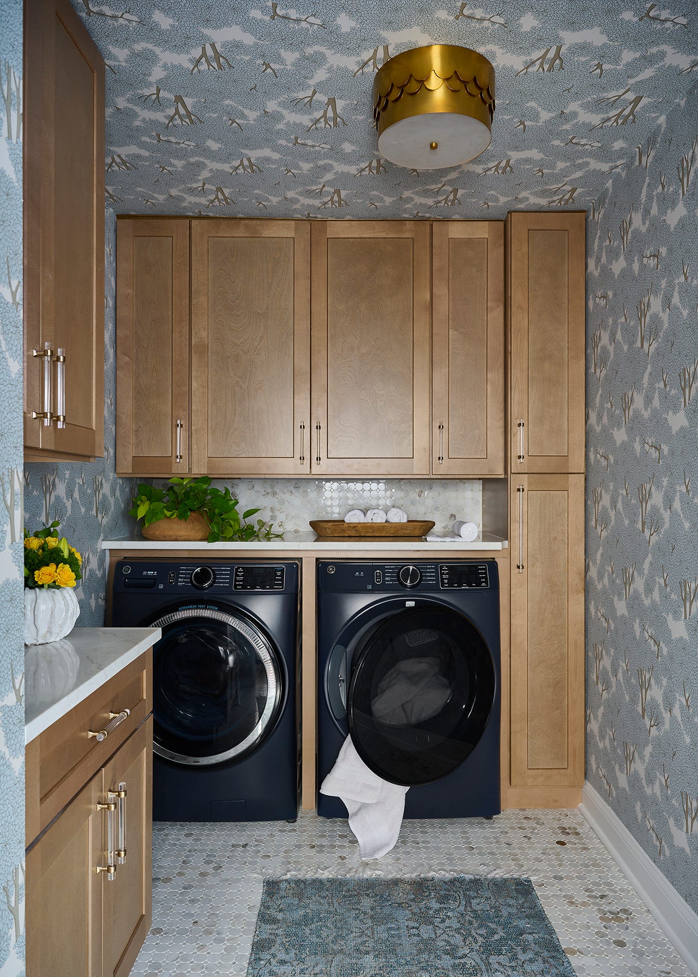 A laundry room features light wood cabinets, front-loading washing machines, and blue tree-patterned wallpaper covering the walls and ceiling.