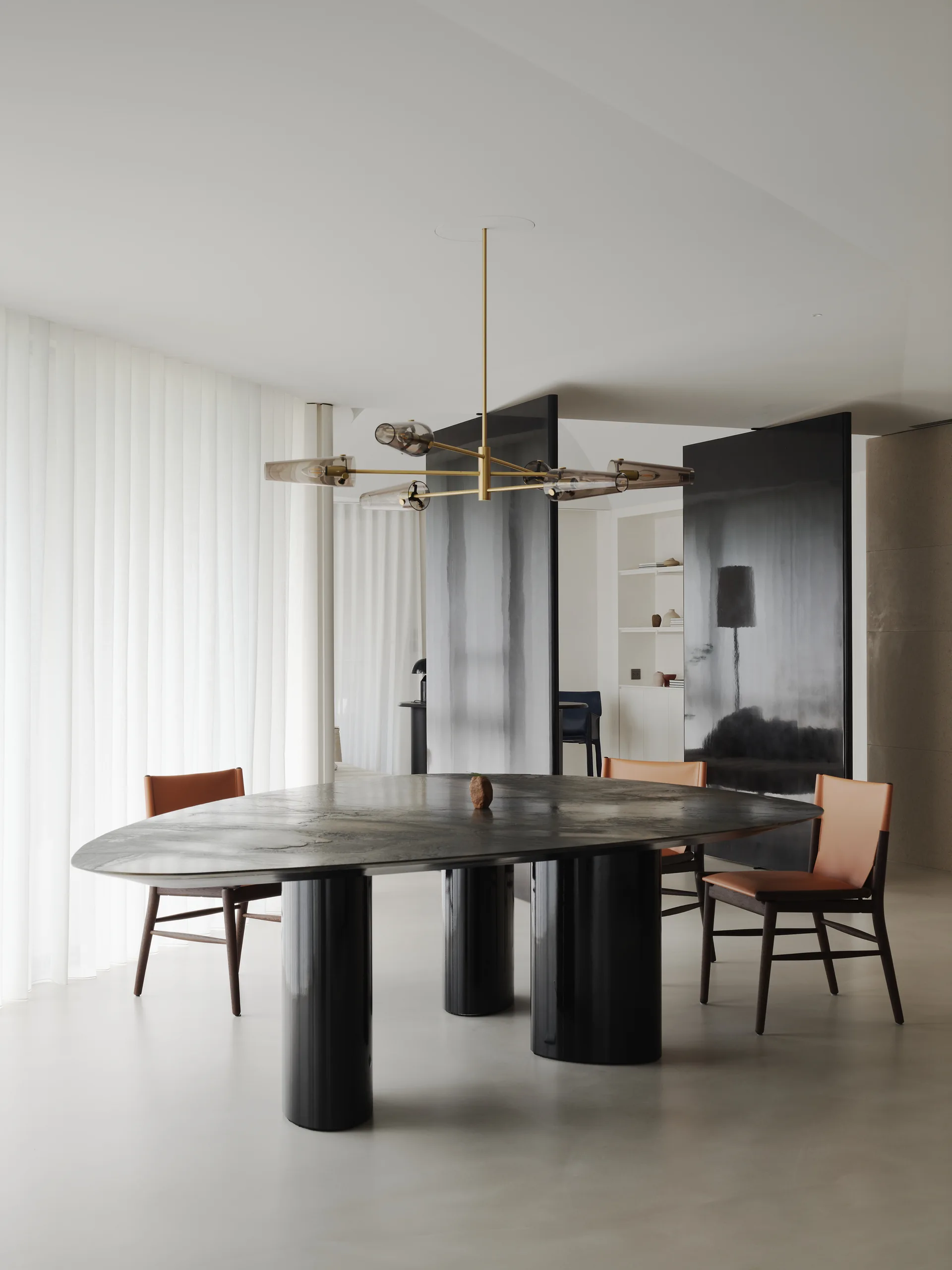 An oval dark wood dining table with glossy black pedestal bases sits on a light floor, surrounded by tan leather chairs under a brass chandelier.