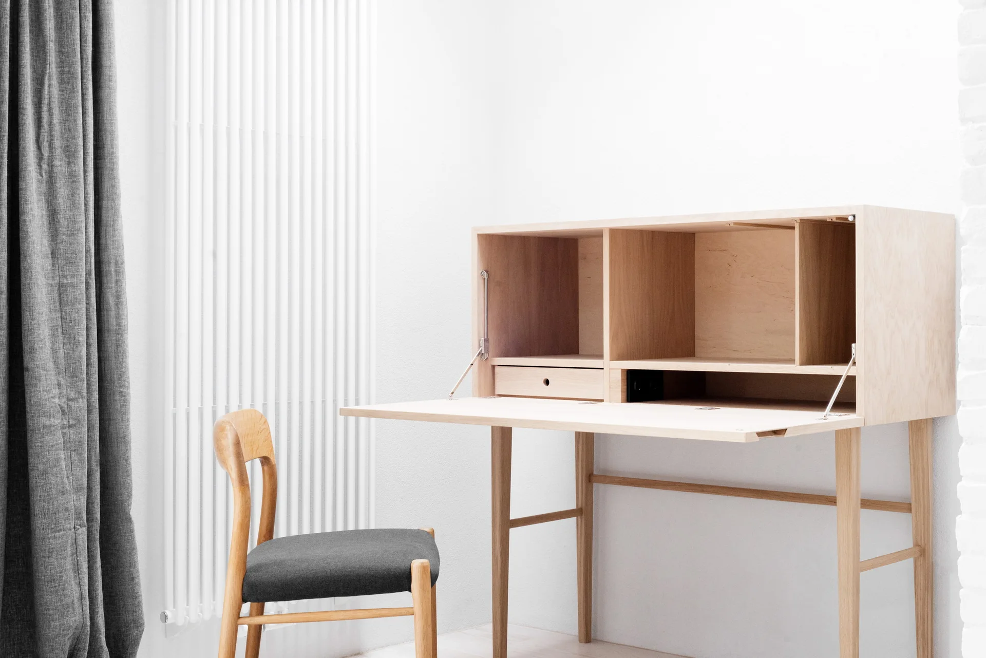 A light wood secretary desk with an open fold-down lid stands next to a wooden chair with a grey cushion against a white wall.