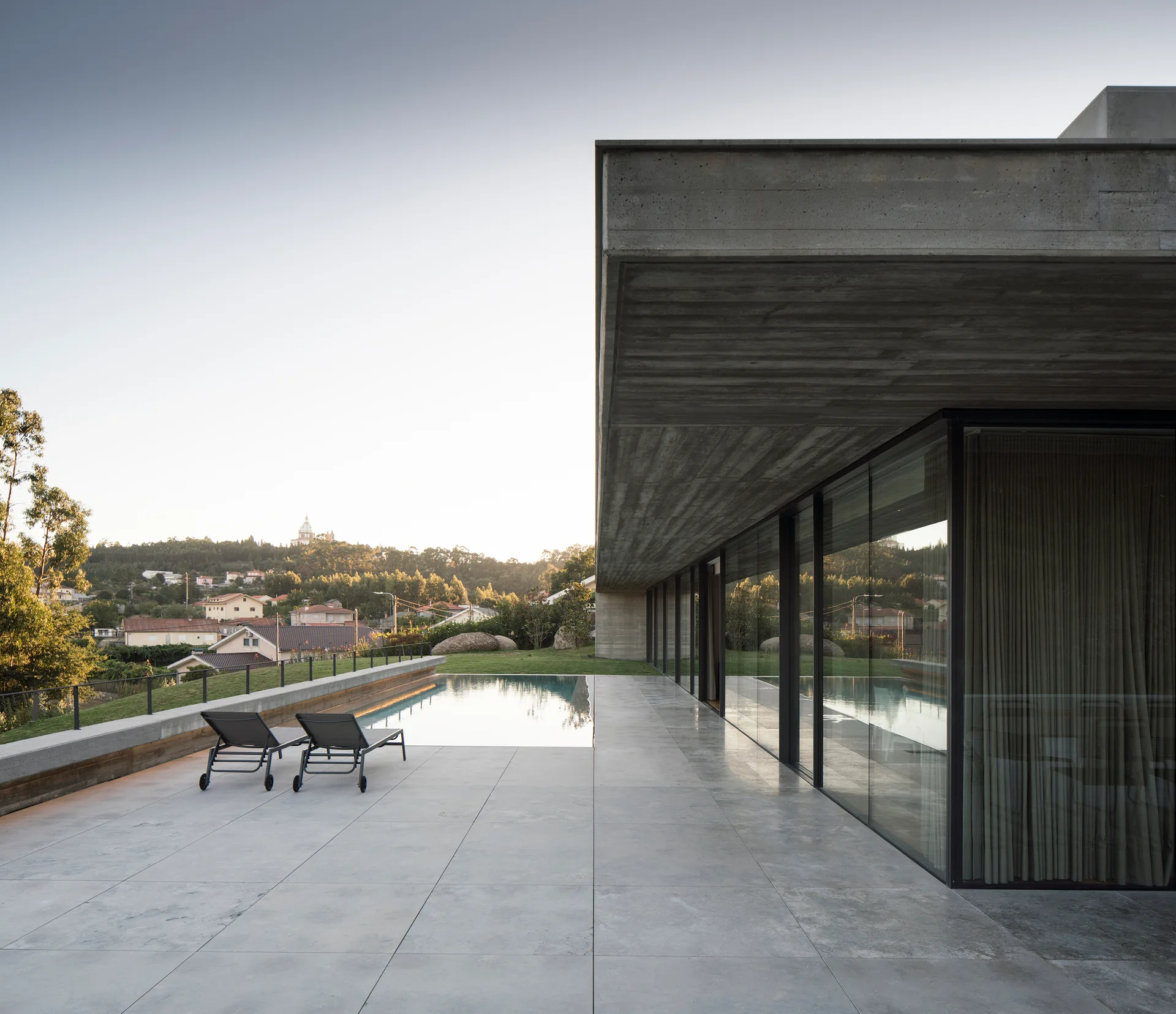 A minimalist concrete patio with two black sun loungers beside a swimming pool under a board-formed concrete roof overhang.