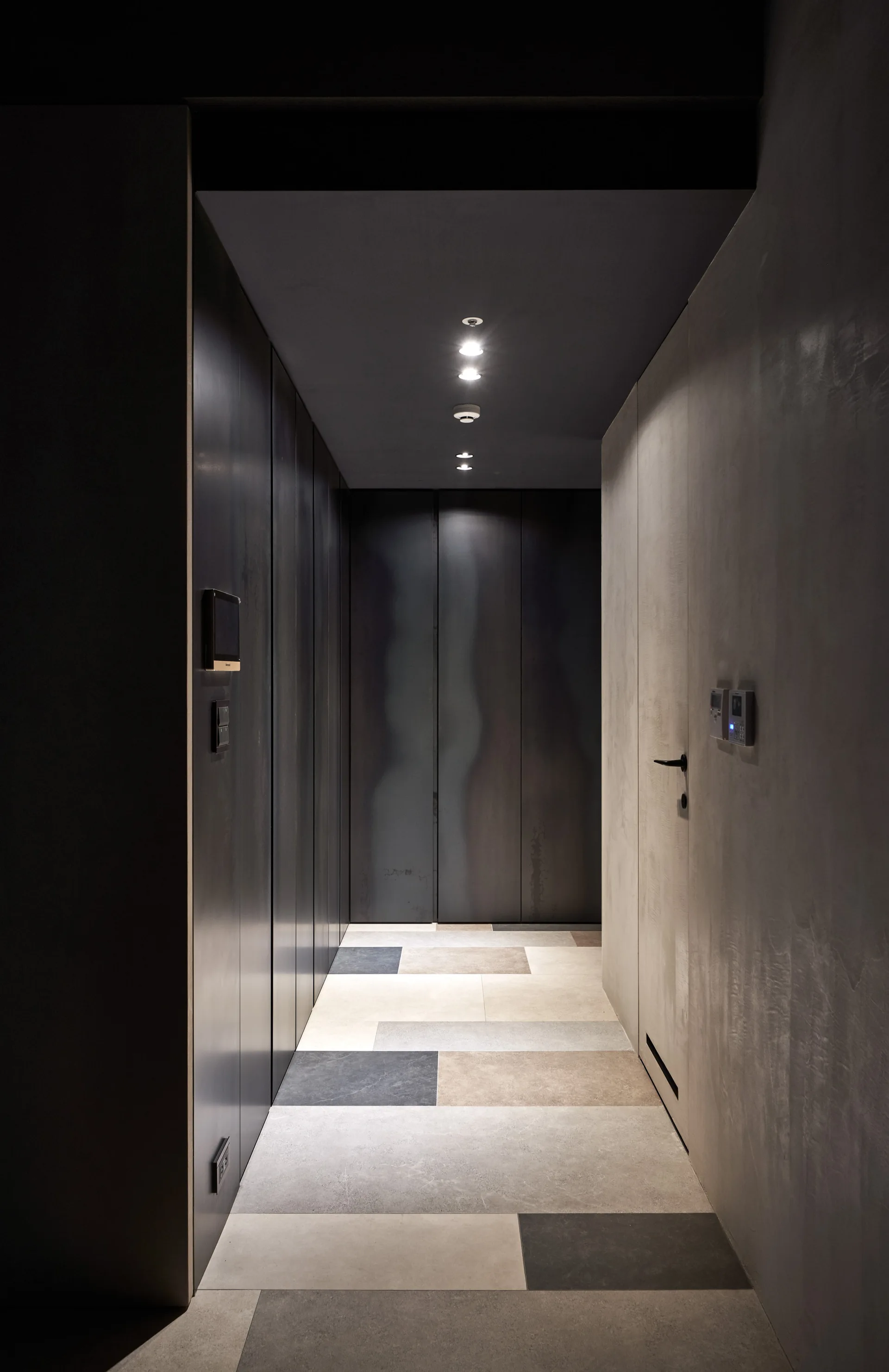 A narrow minimalist hallway features dark charcoal wall panels, a gray textured plaster wall, and large rectangular floor tiles in neutral tones.