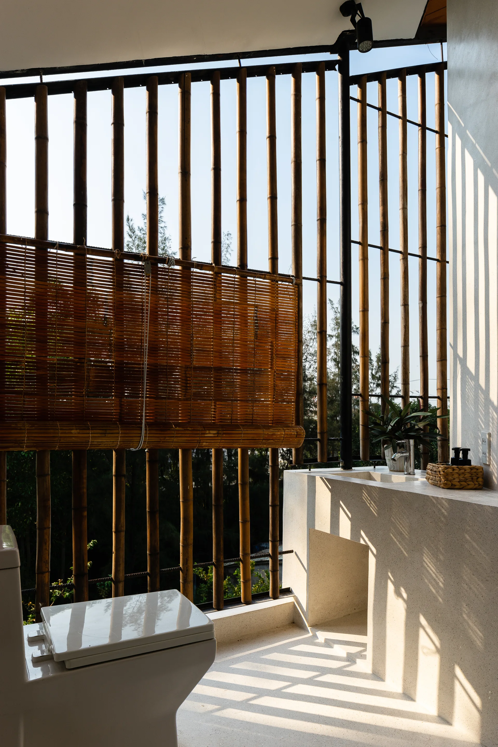 An open-air bathroom features a white concrete vanity and toilet set against a vertical bamboo screen with sunlight filtering through.