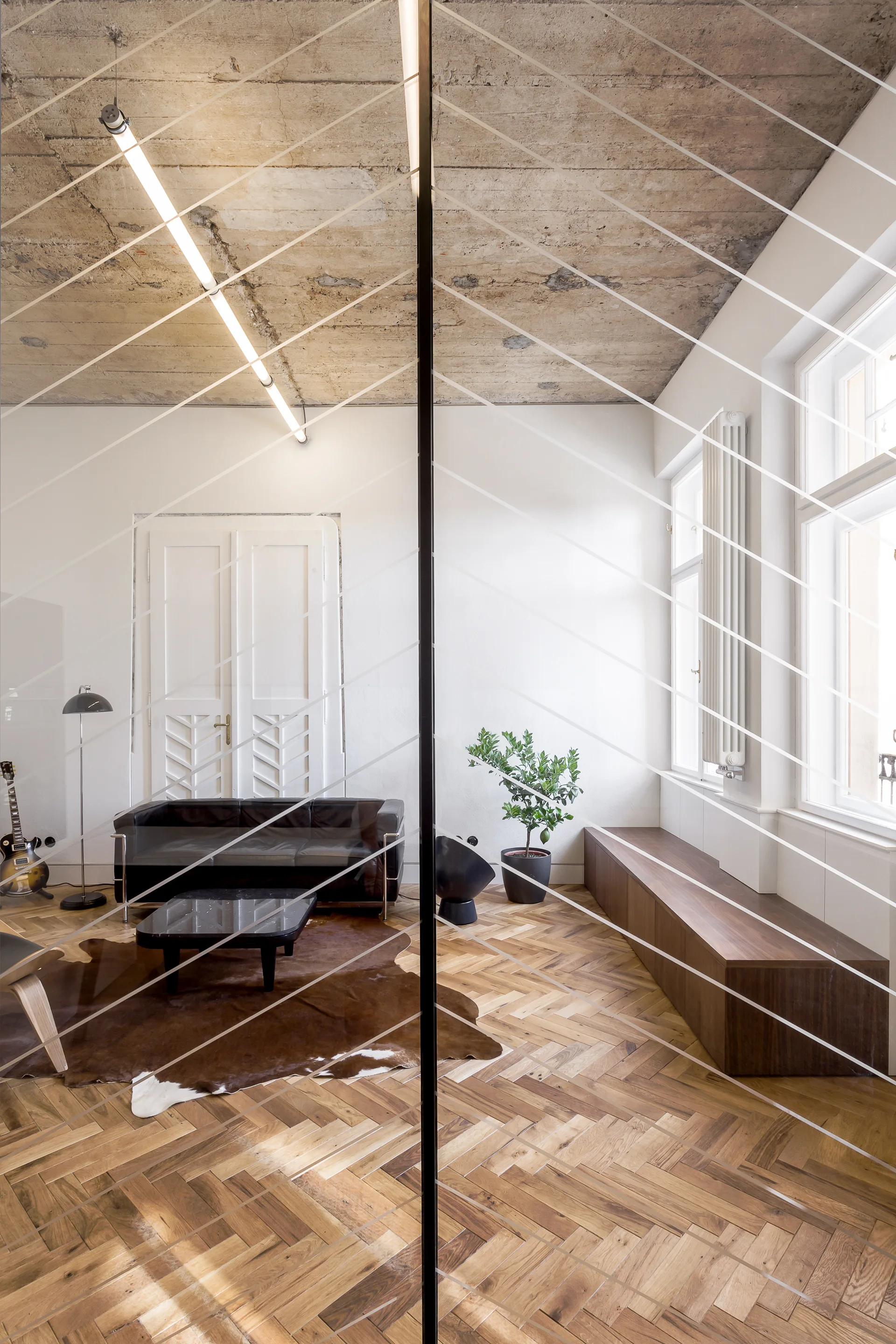 A contemporary living room viewed through glass, featuring a raw concrete ceiling, herringbone wood floors, a black leather sofa, and a long wooden bench.