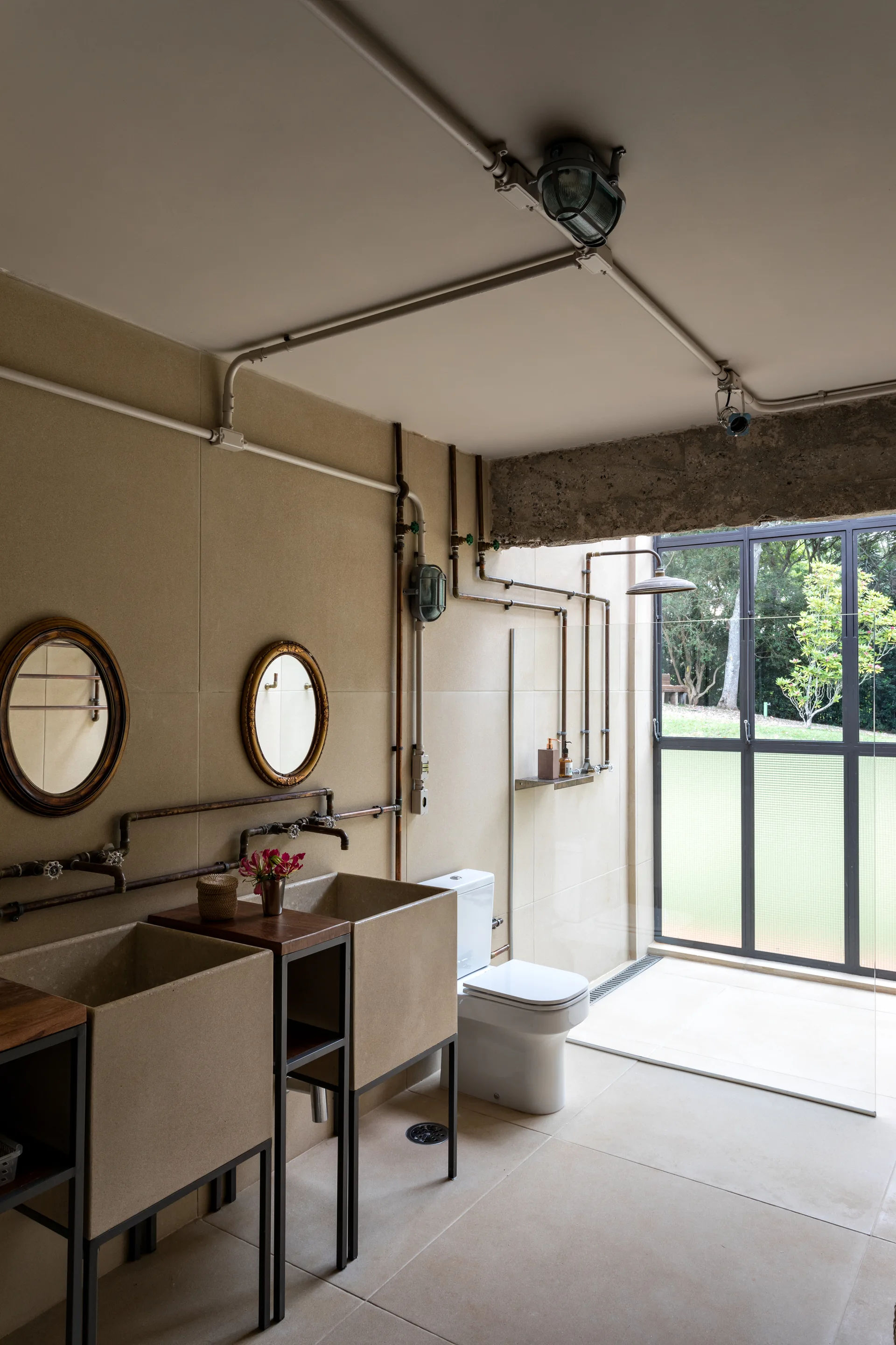 An industrial bathroom features two rectangular stone basin sinks, oval mirrors, exposed copper pipes, and a glass door opening to a green yard.