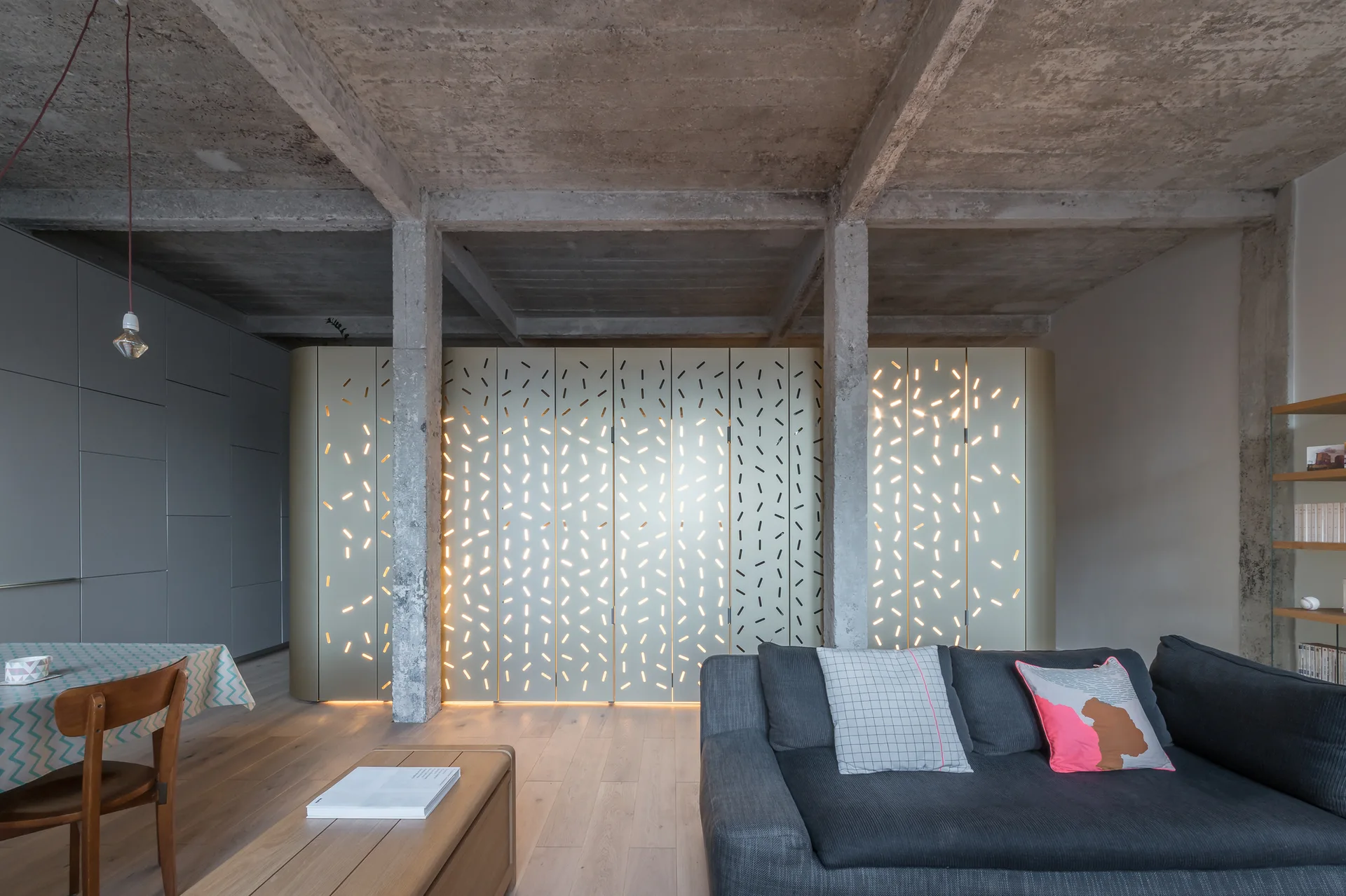 A modern living room features a dark grey sofa, light wood flooring, concrete ceiling beams, and a backlit perforated room divider panel.
