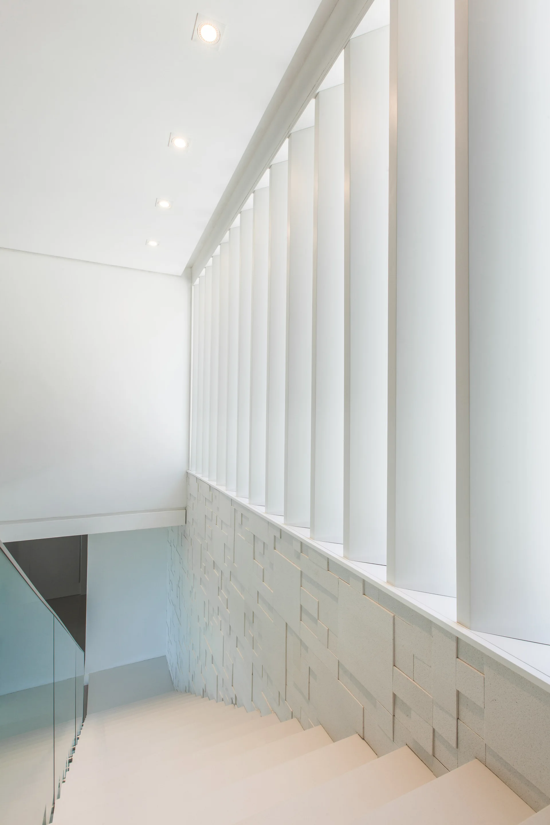 A modern white staircase with a geometric textured wall, glass railing, and high vertical louvered panels under recessed lighting.
