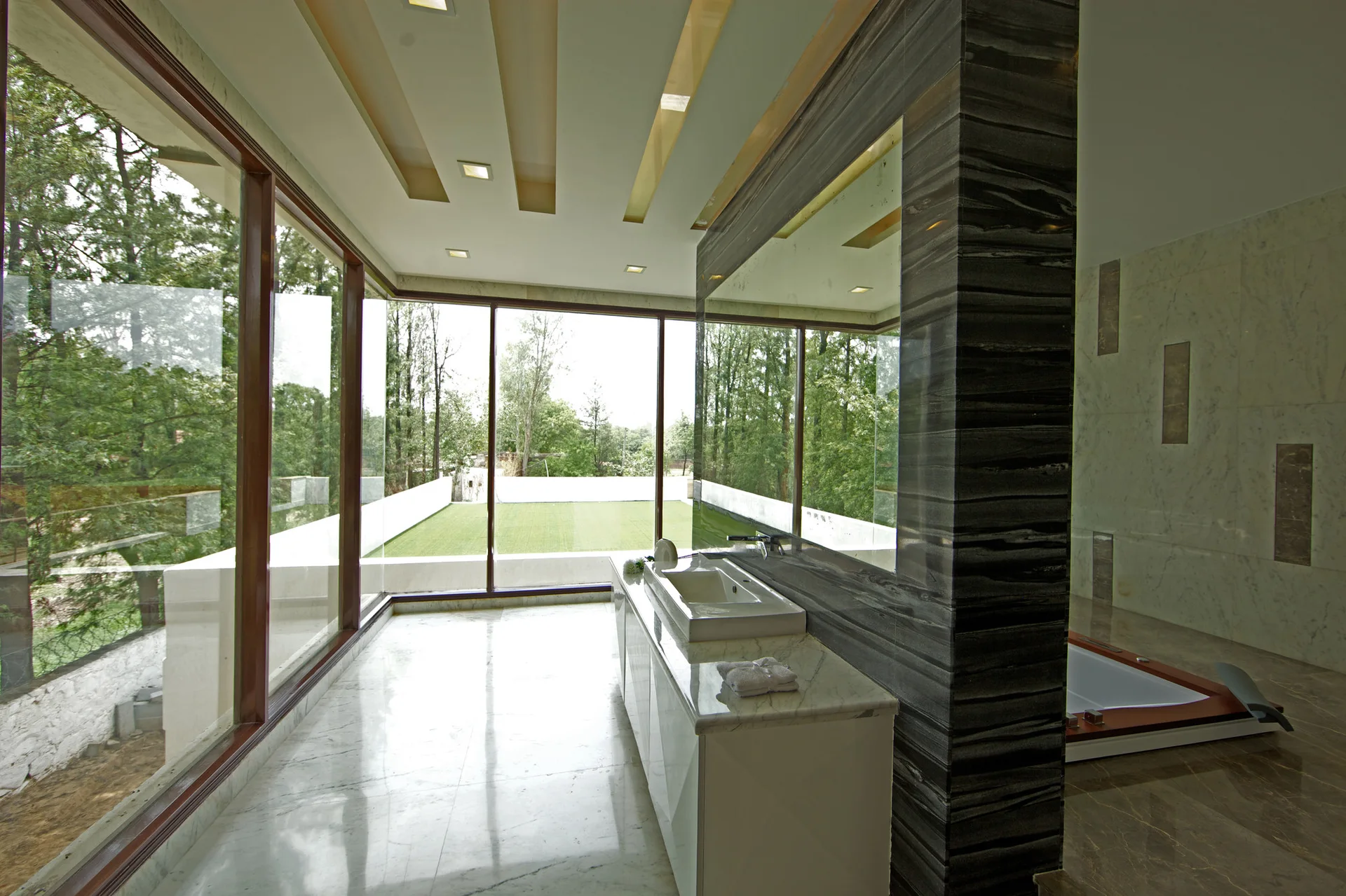 A contemporary bathroom features a dark veined stone partition wall, a white vanity with sink, a large mirror, and floor-to-ceiling windows.