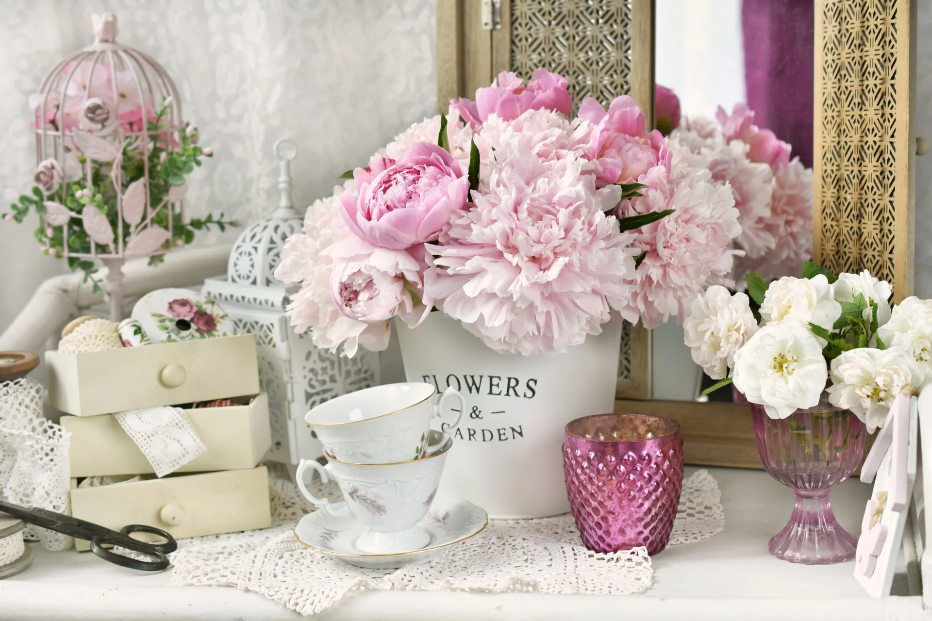 A tabletop display features a bucket of pink peonies, stacked porcelain teacups on a lace doily, and a wooden decorative mirror.