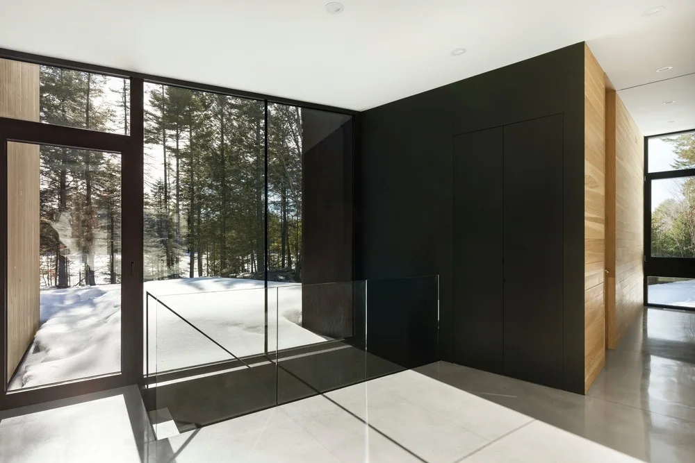 A minimalist hallway features polished concrete floors, natural wood wall paneling, and a frameless glass staircase railing next to large windows.