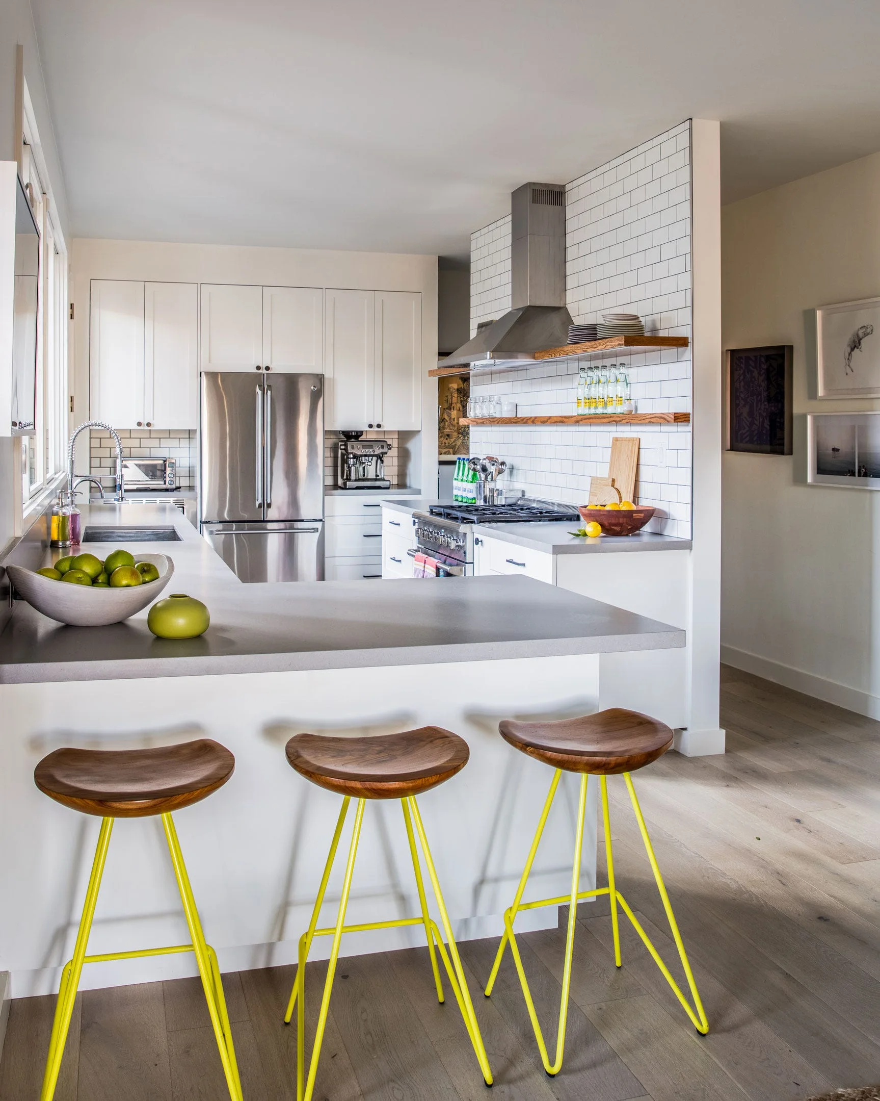 A modern white kitchen features grey countertops, white subway tiles, wooden floating shelves, and three bar stools with bright yellow metal legs.