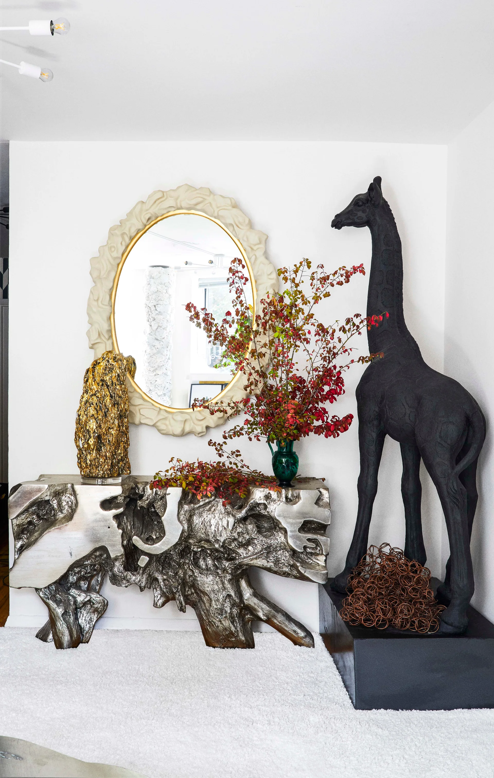 A sculptural silver console table topped with a gold object and branches, situated next to a large black giraffe sculpture and beneath an oval mirror.