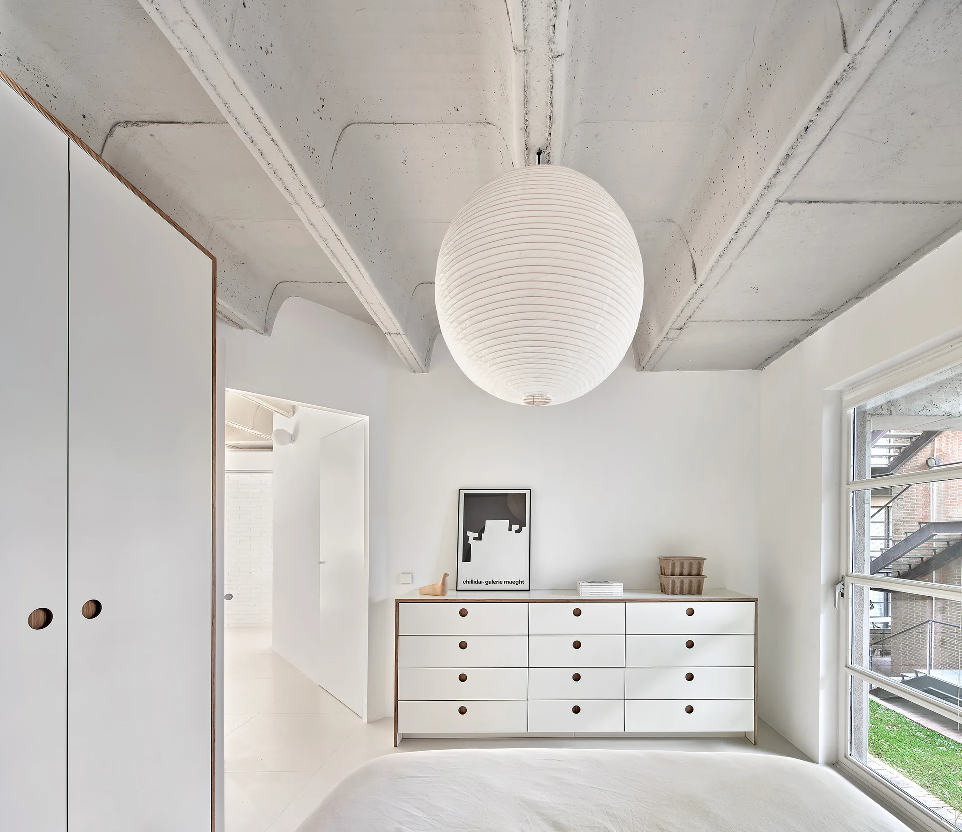 A minimalist white bedroom features an exposed concrete ceiling, a large spherical paper lantern pendant, and a white dresser with circular wooden pulls.