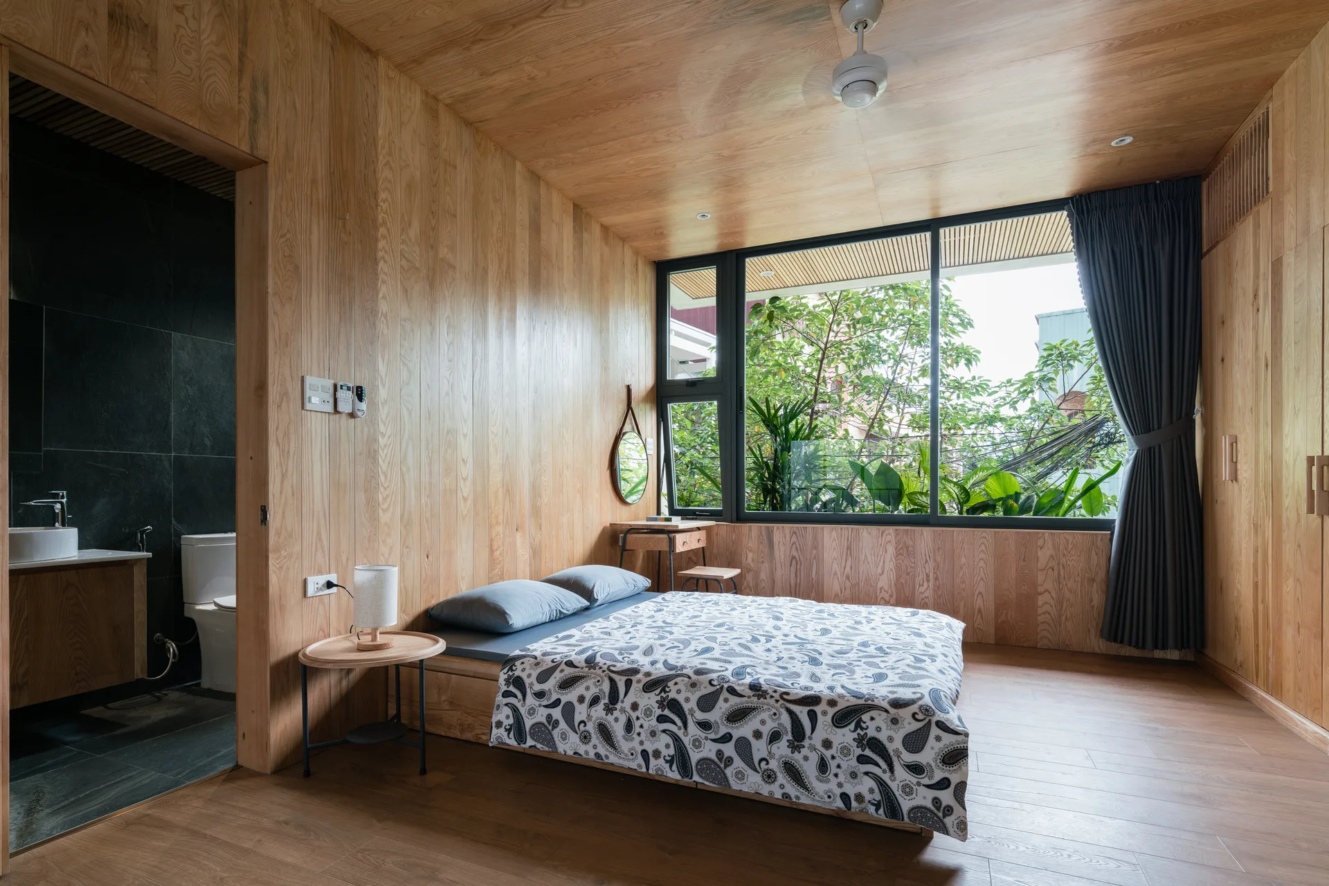 A minimalist bedroom featuring light wood paneled walls and ceiling, a low bed with patterned bedding, and a large window showcasing lush green trees.