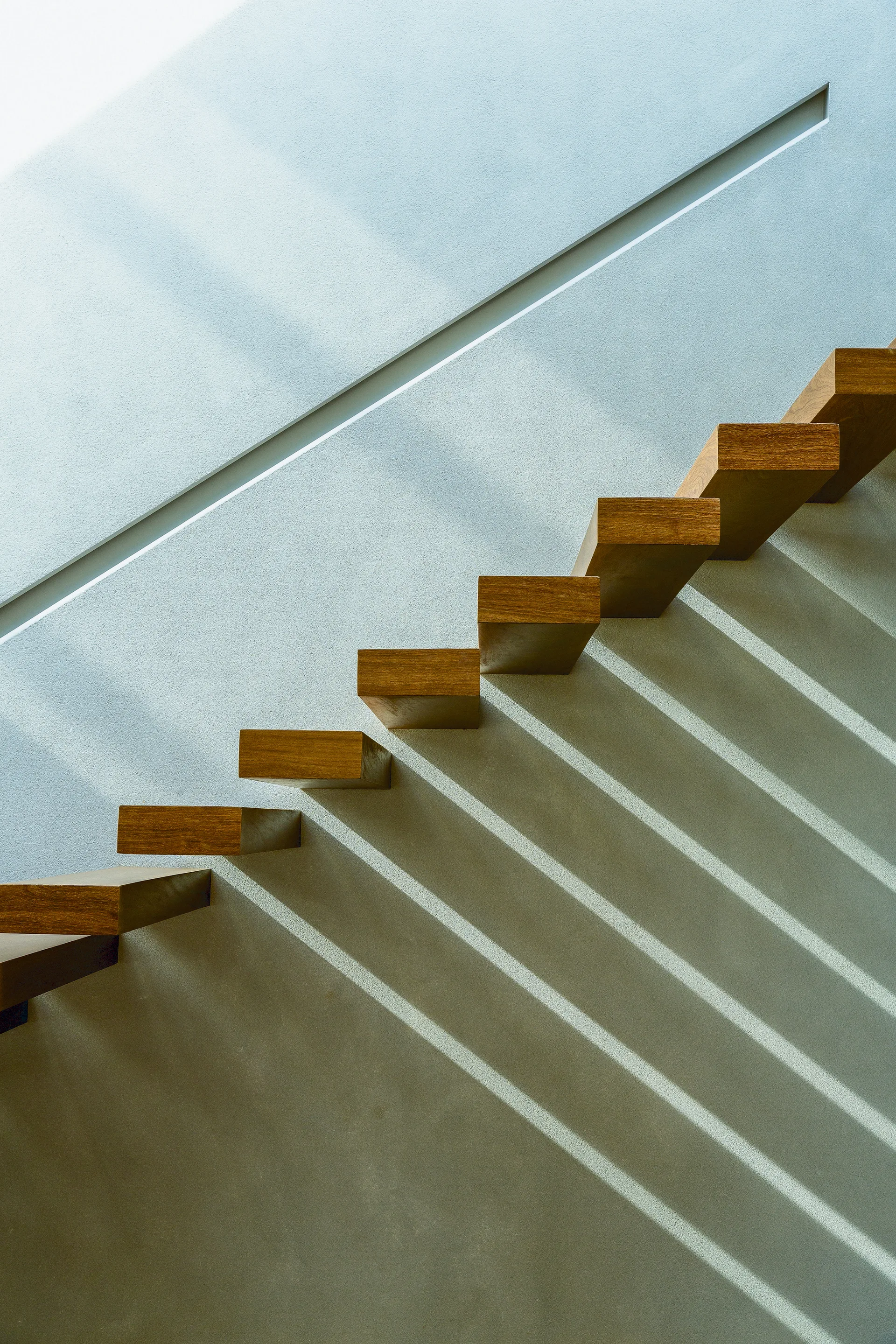 Minimalist floating wooden stair treads are mounted on a smooth gray wall with a diagonal recessed channel.