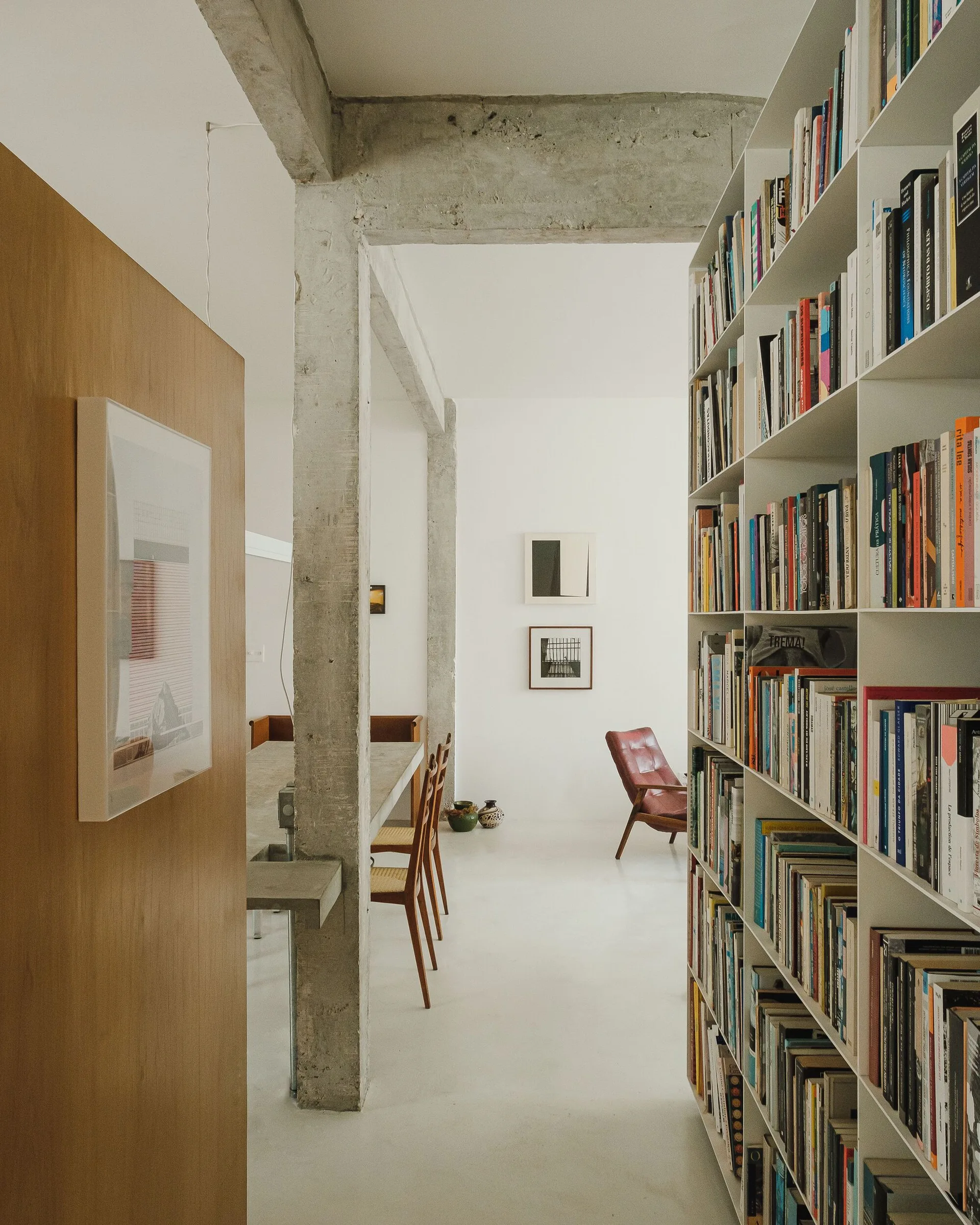 An open plan interior features a tall white floor-to-ceiling bookshelf filled with books, an exposed concrete pillar, and a wood-paneled wall.