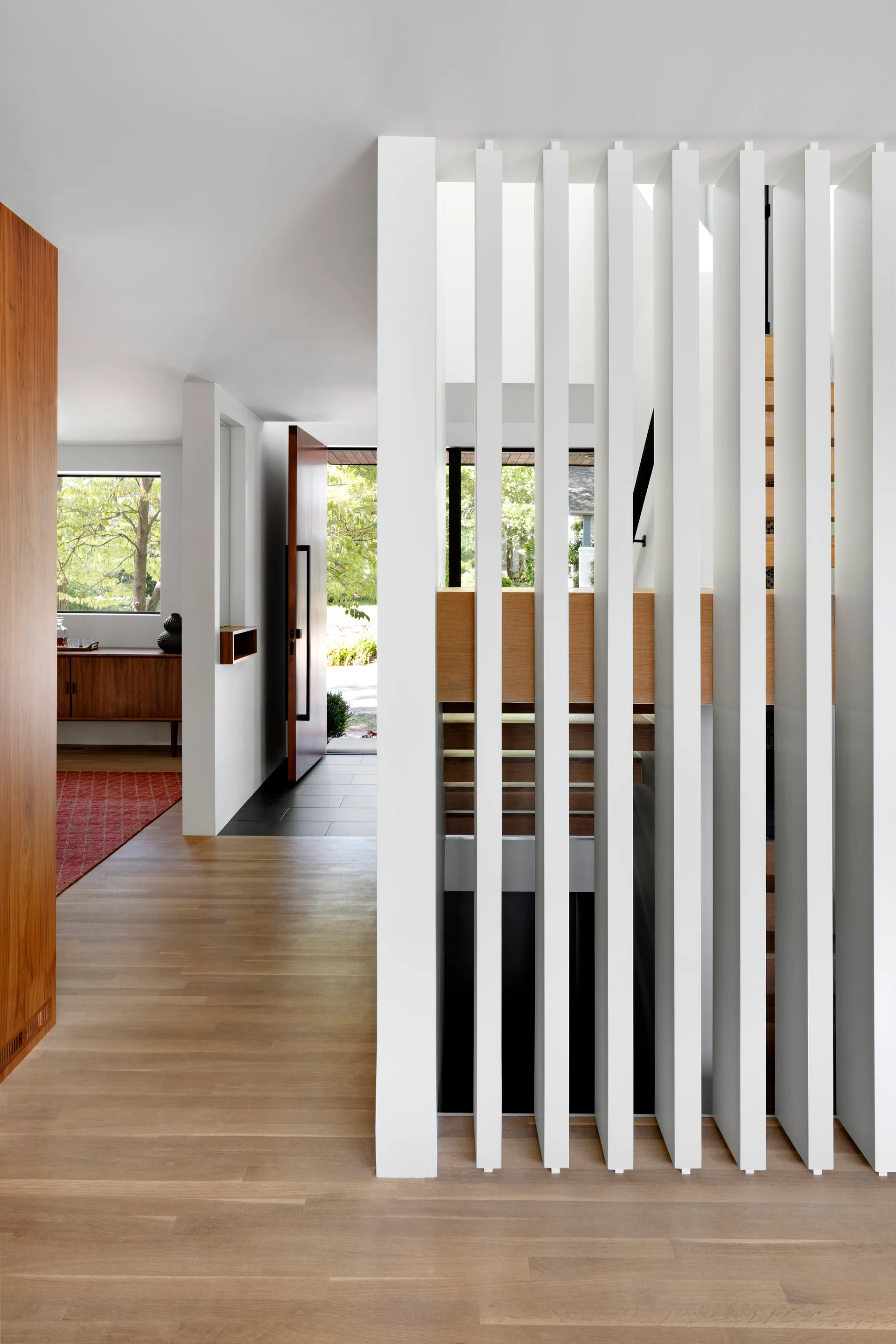 A minimalist entryway features light hardwood floors, an open wood front door, and a white vertical slat partition screening a wooden staircase.