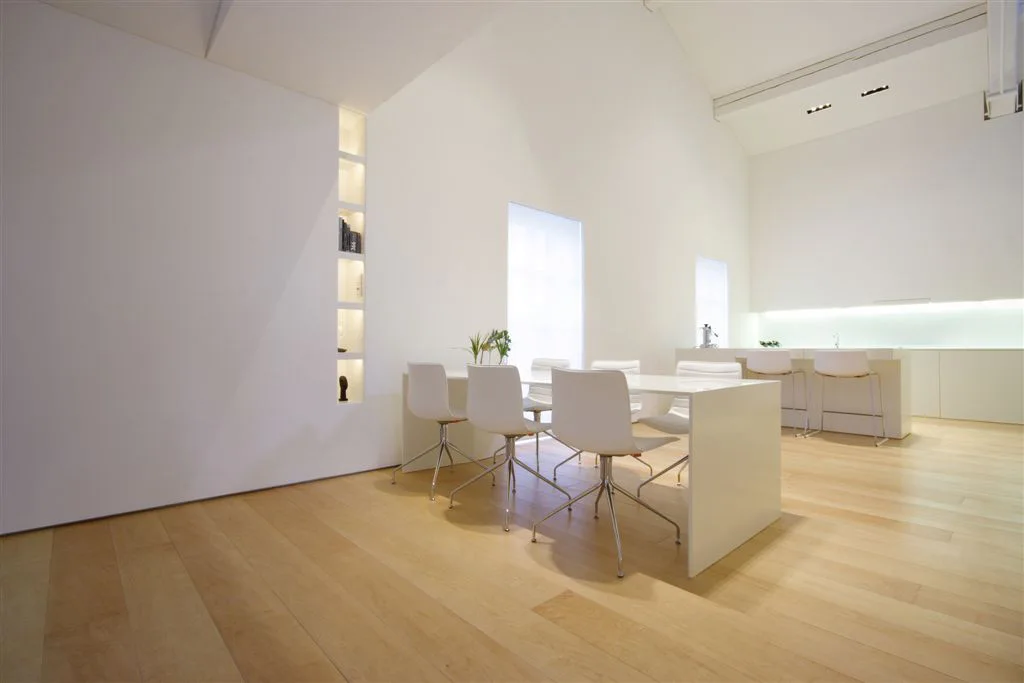 A minimalist white dining table and chairs sit on light wood flooring next to a vertical recessed wall shelf.