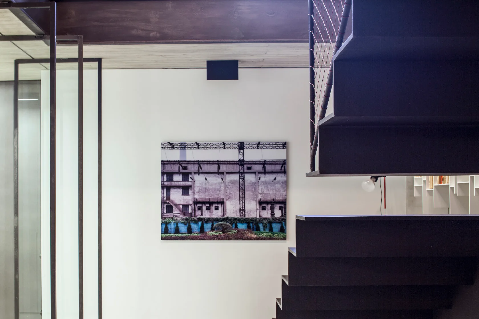 A minimalist dark metal staircase frames a white wall displaying a framed print of an industrial factory landscape.