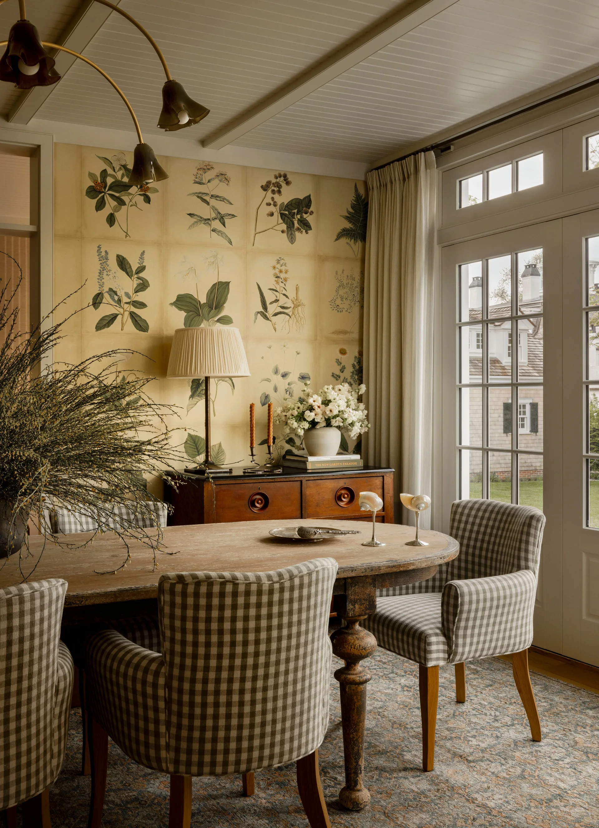A wooden dining table and gingham-patterned chairs set against a yellow botanical wallpaper wall next to a glass French door.