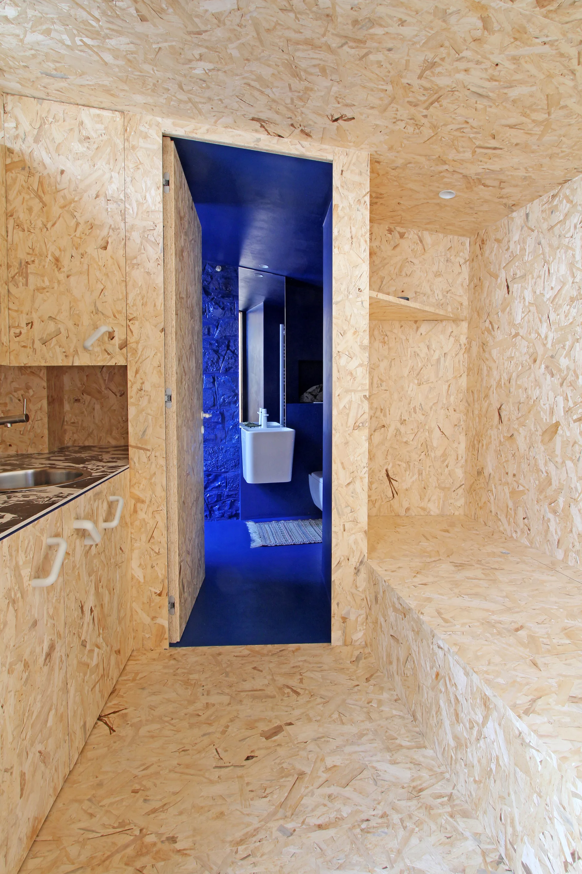 A small kitchen finished entirely in oriented strand board paneling features an open doorway leading into a vibrant blue bathroom.
