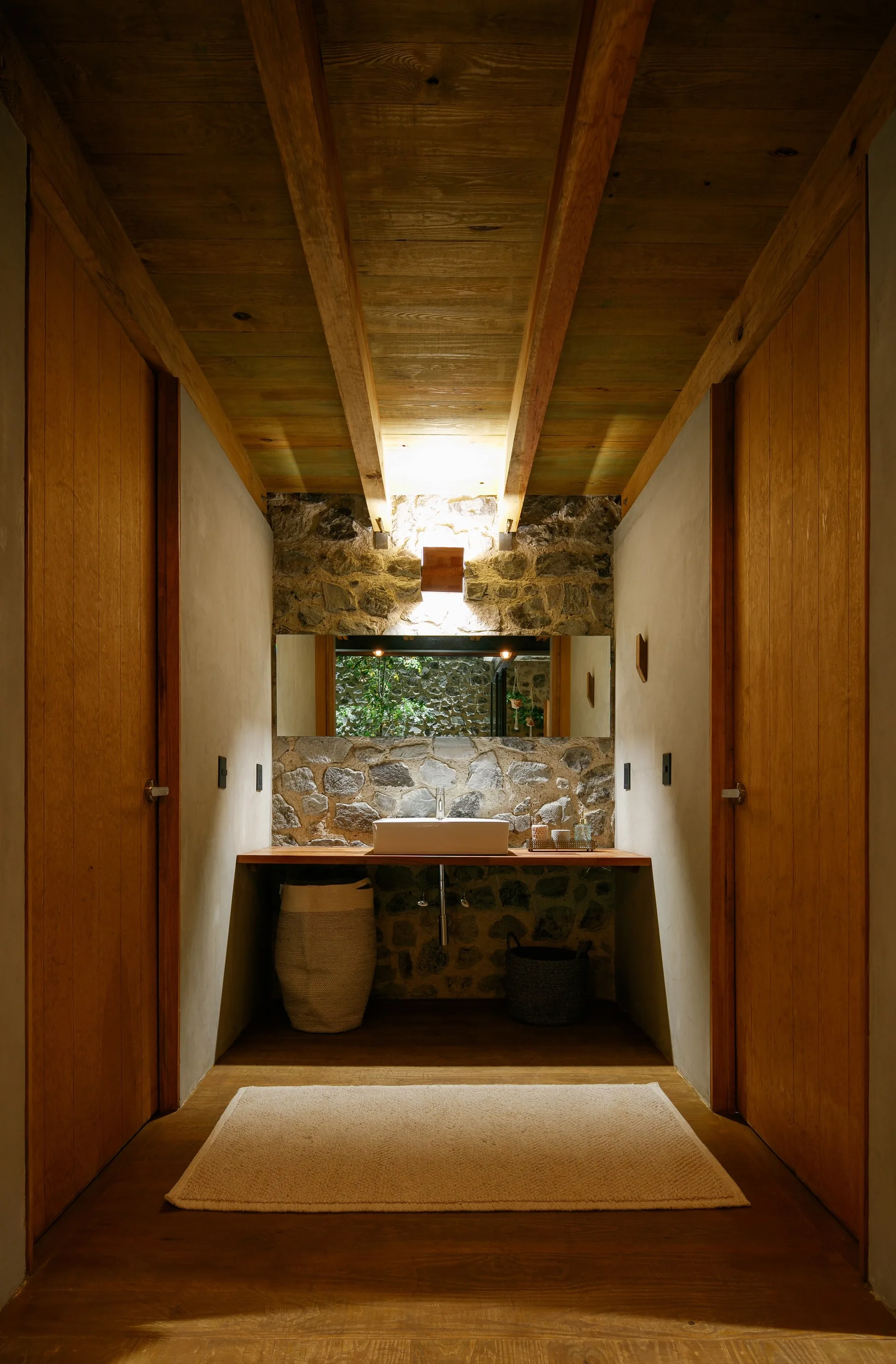 A rustic bathroom features a floating wooden vanity with a white vessel sink against a natural stone wall under an exposed timber ceiling.