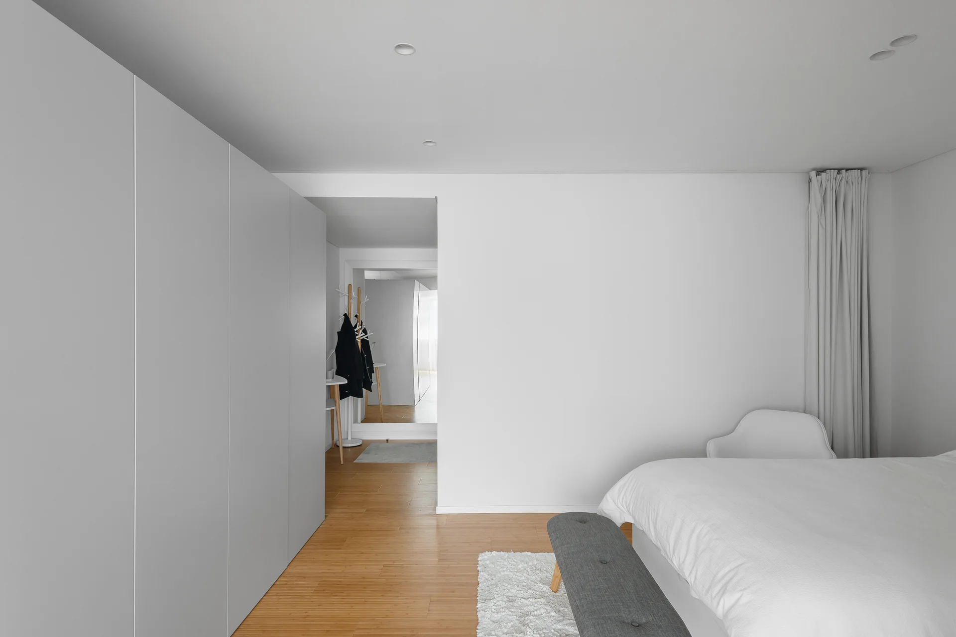 A minimalist white bedroom featuring a large built-in wardrobe, a grey upholstered bench on a shag rug, and light wood floors.
