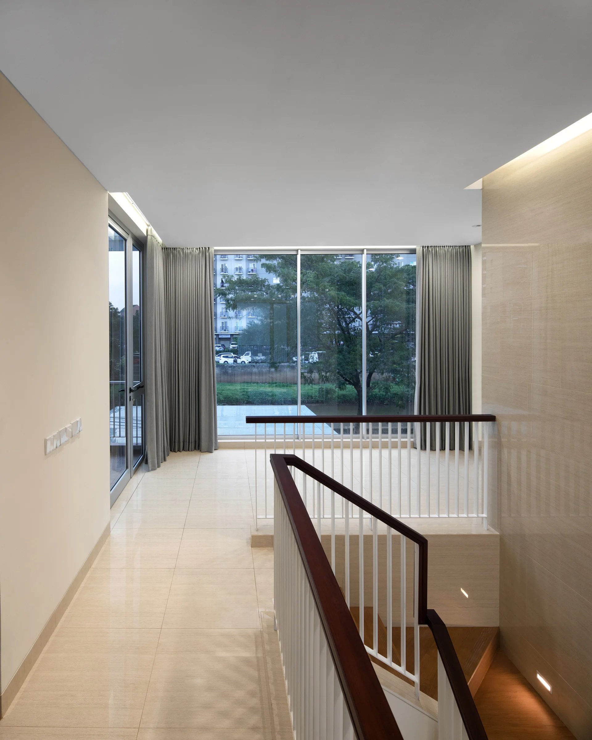 A modern staircase landing features glossy beige tiled flooring, a wooden handrail with white spindles, and large floor-to-ceiling windows.