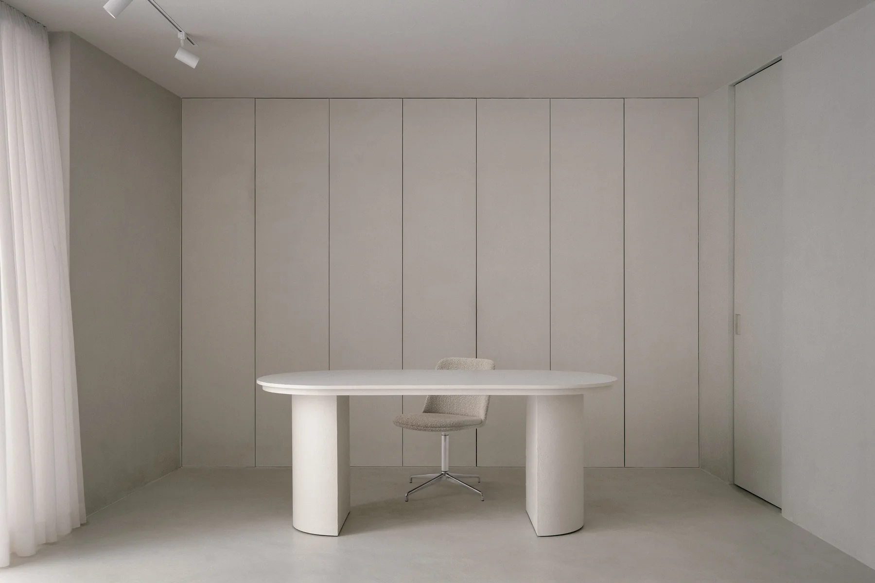 A minimalist beige home office features an oval pedestal desk and a boucle swivel chair set against floor-to-ceiling vertical wall paneling.